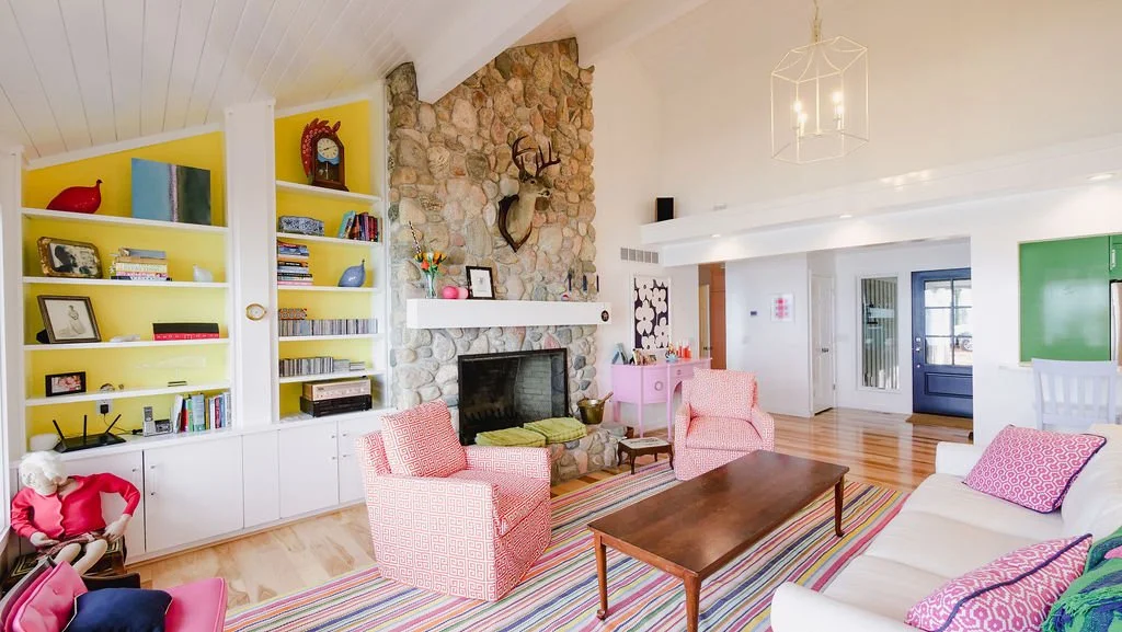 Living room with a stone fireplace, pink and white armchairs, a white sofa with pink pillows, a wooden coffee table, colorful striped rug, built-in yellow shelves with decorative items and books, and a white ceiling with a geometric chandelier.