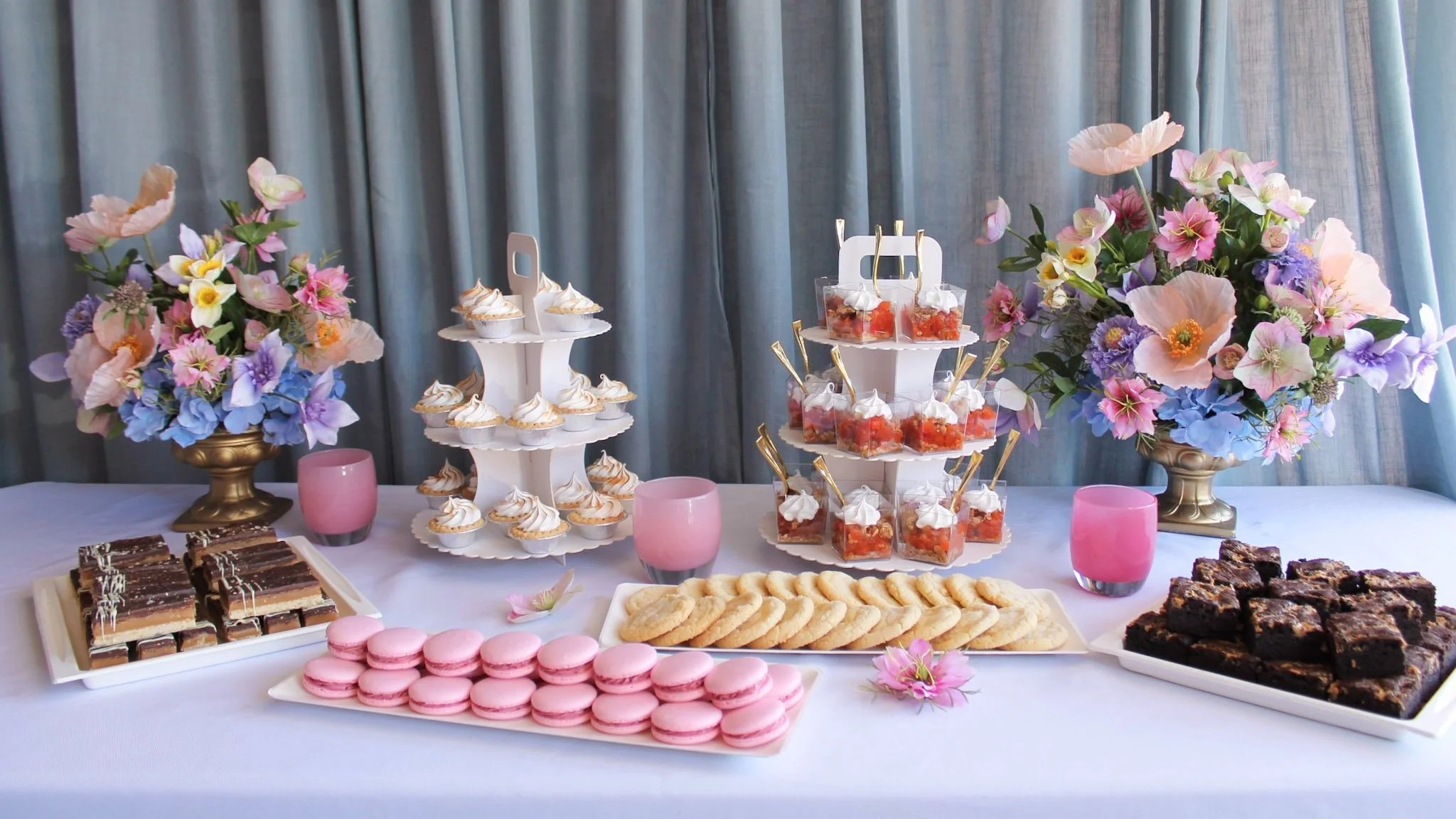 A dessert table with pink macarons, layered bars, and brownies, decorated with large pink and purple flowers in vases.