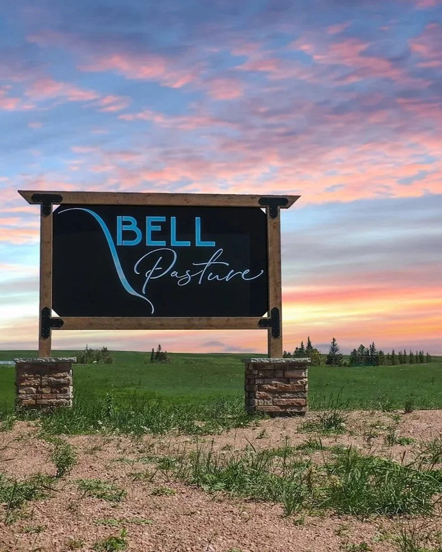 A sign for Bell Pasture in a rural area with a grassy field and trees, under a colorful sunset sky.