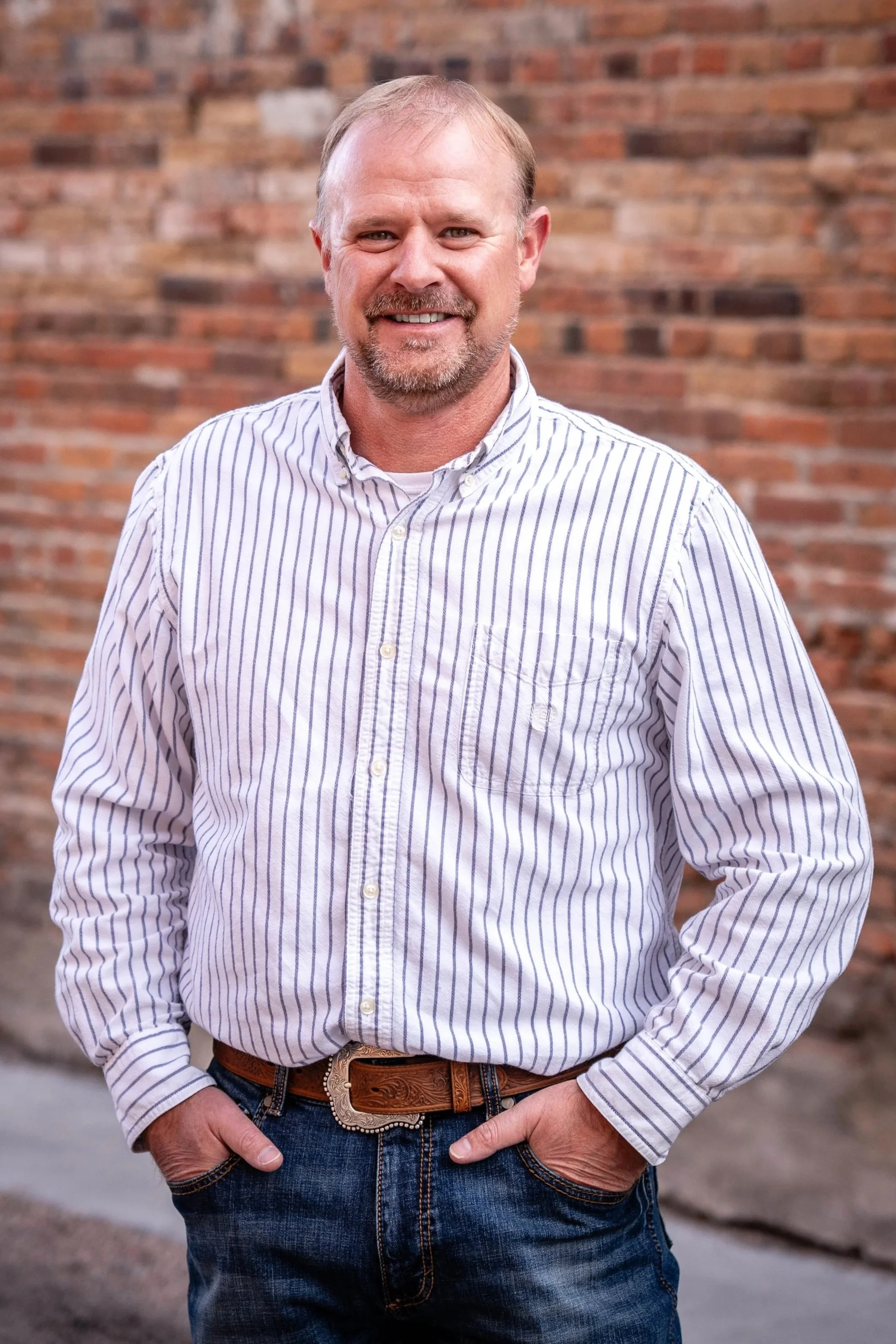 A man with light hair, beard, and mustache wearing a white and blue striped button-up shirt, blue jeans, and a brown leather belt stands in front of a brick wall.