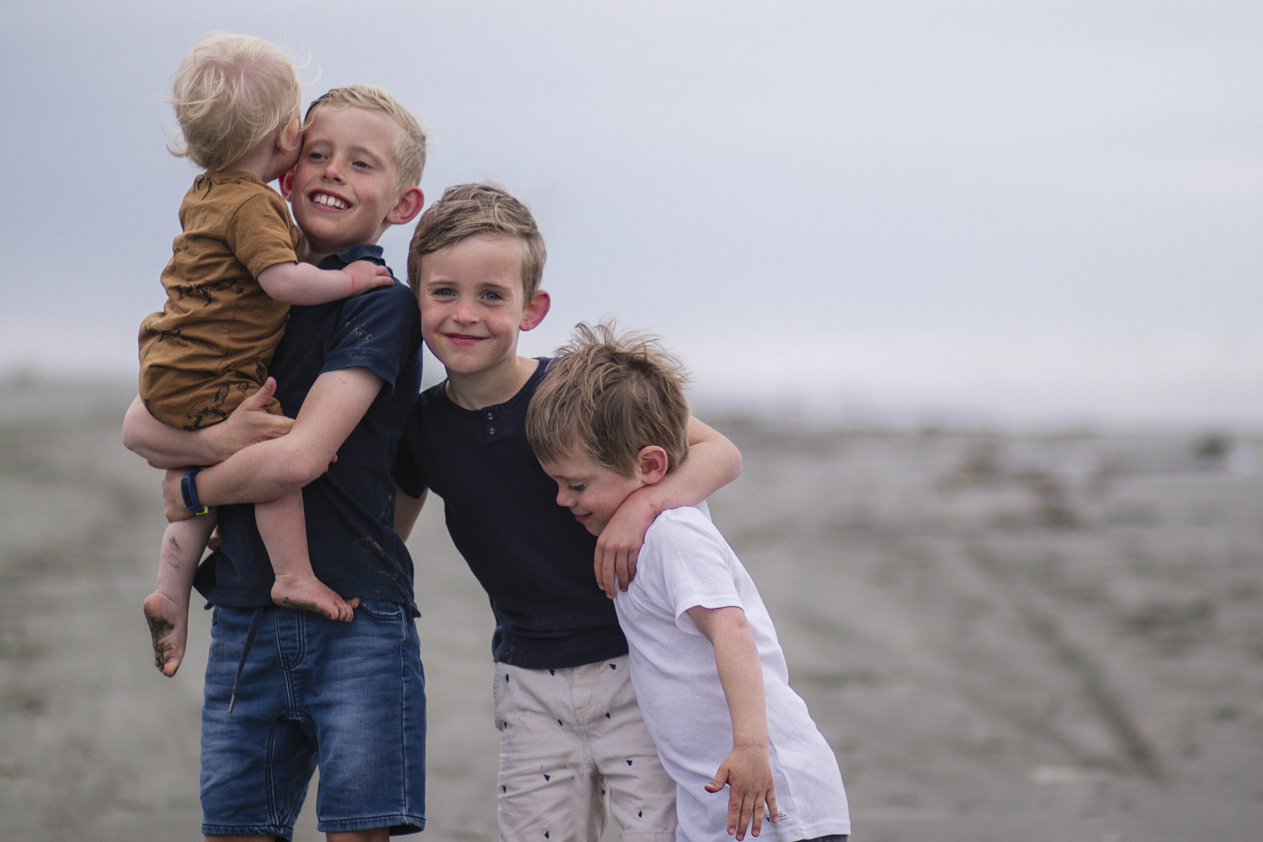 Four brothers on a beach, hugging and playing, with the ocean in the background.