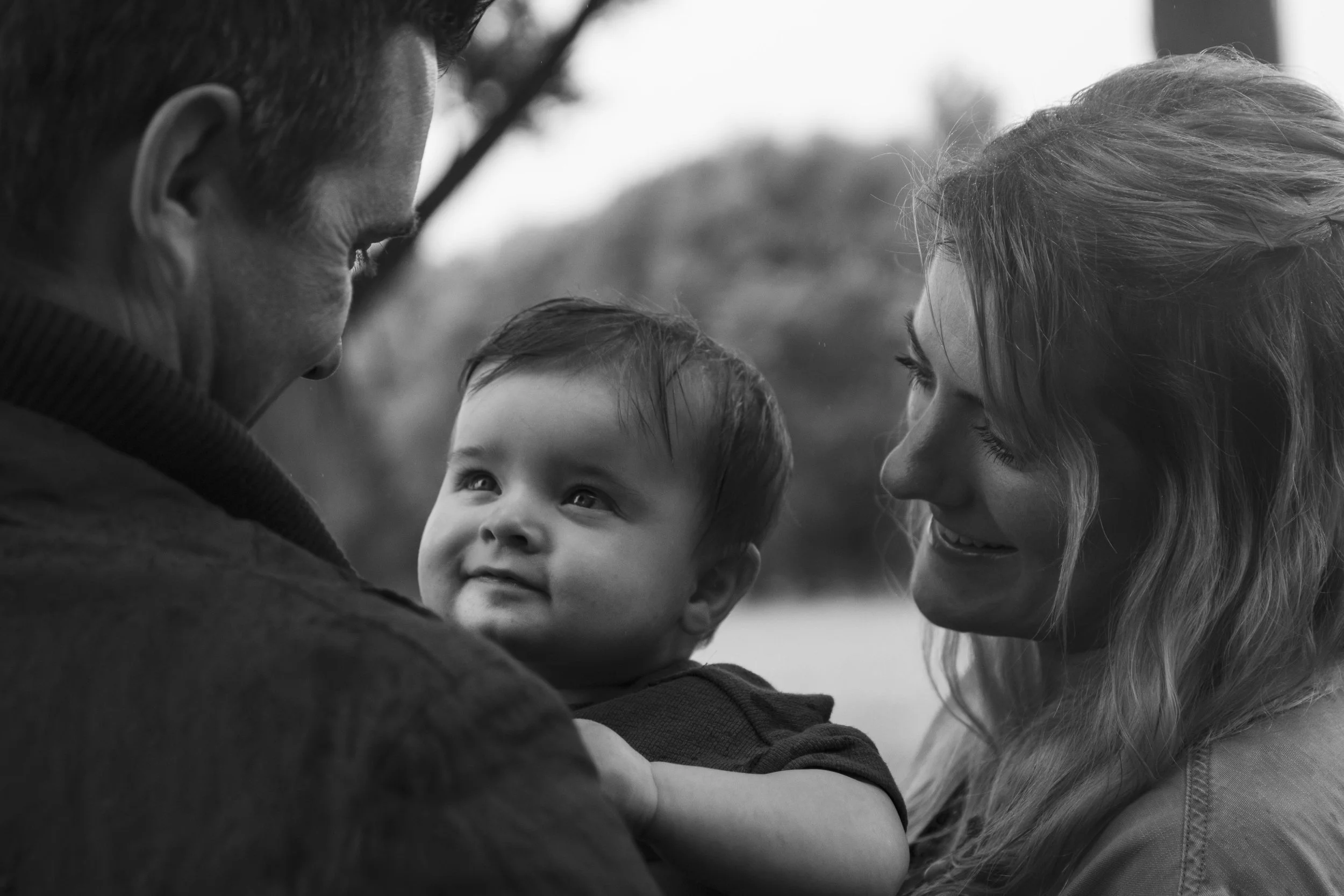A black and white photograph of a family showing a man, woman, and young child outdoors, with the child being held by the man. The family appears happy and connected.