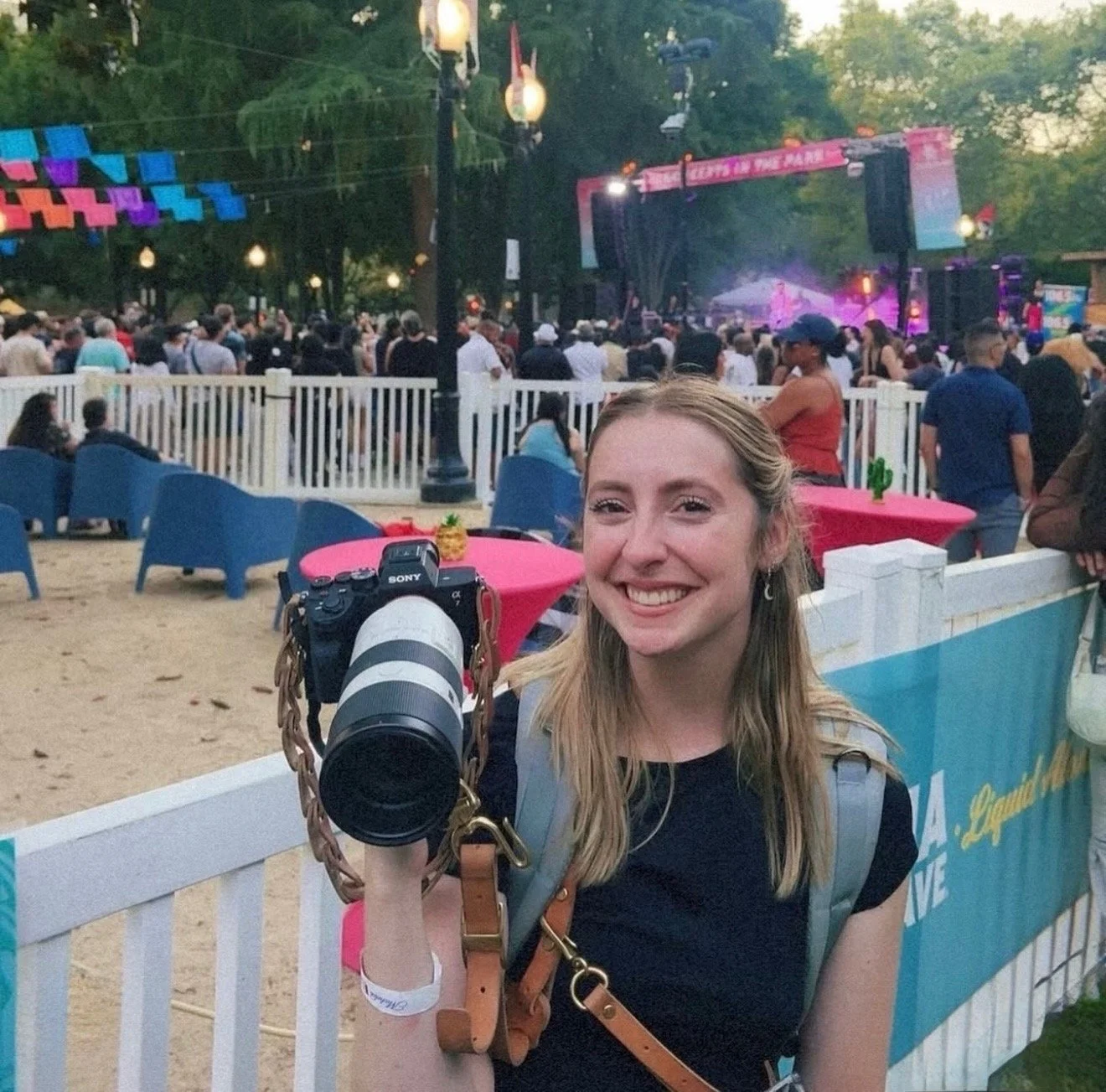 A smiling young woman with a camera hangs around her neck, at an outdoor concert or festival. She has long blonde hair, wears a black shirt, and a wristband. Behind her, a large crowd is gathered in front of a stage, with colorful decorations hanging