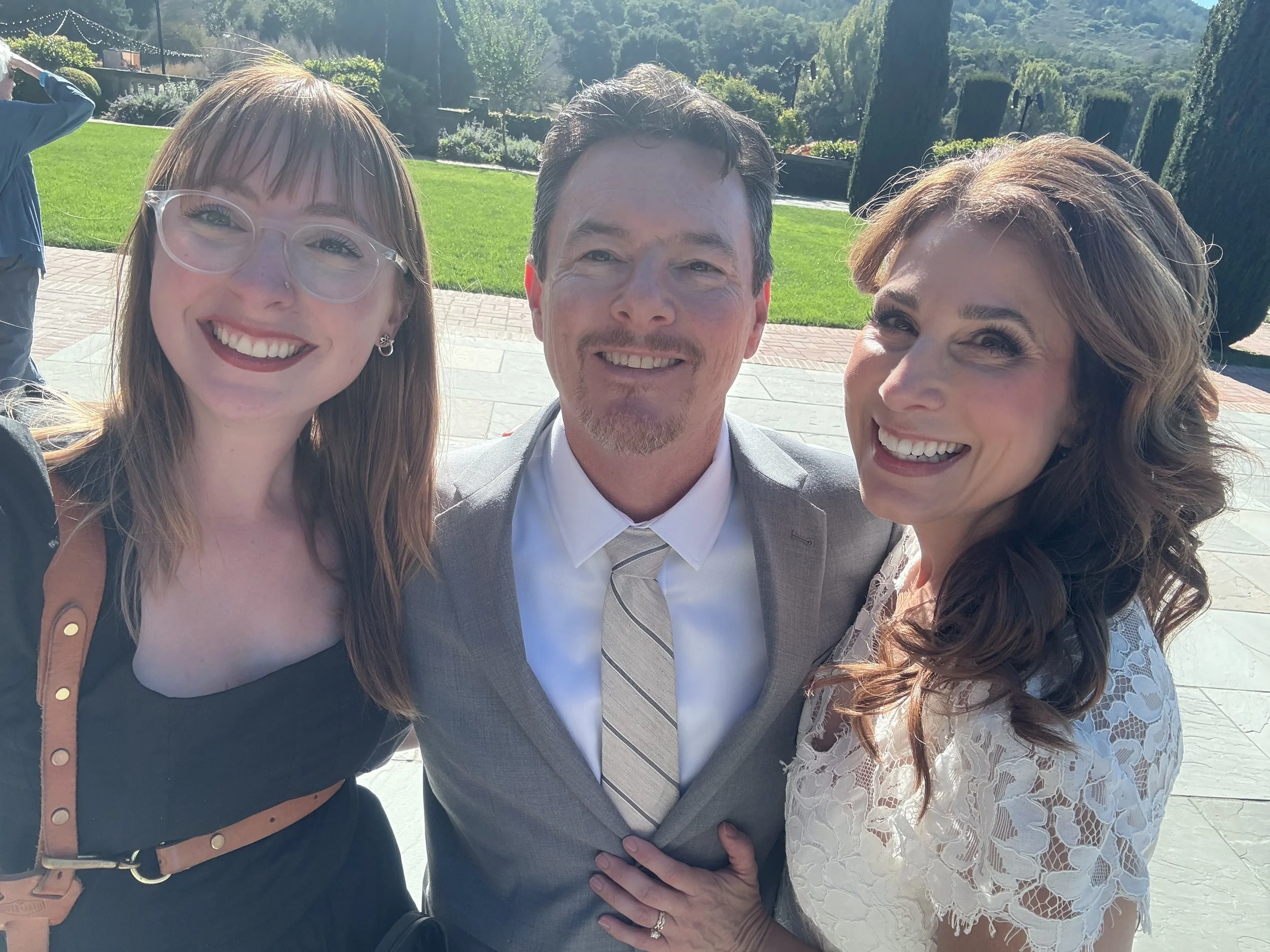 Three people smiling outdoors on a sunny day, with a lush garden background. The woman on the left has long straight hair, glasses, and is wearing a black dress with a tan belt. The man in the middle has short dark hair, a goatee, and is dressed in a