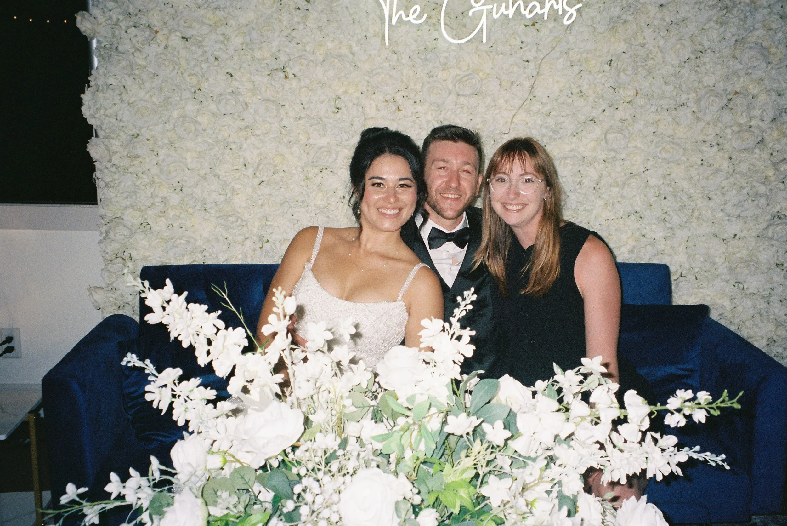 Three people sitting on a blue velvet sofa, partially obscured by a large bouquet of white flowers, smiling at the camera, against a white floral wall with a sign that says 'The Guhars' in cursive.