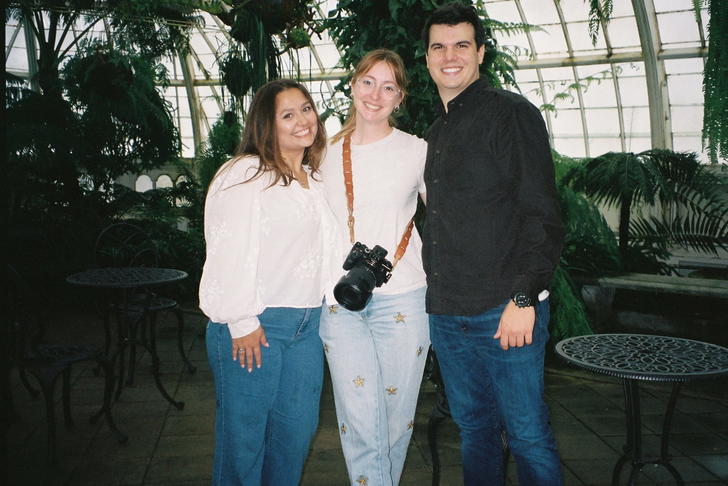 Three people posing together inside a greenhouse. The person in the middle has a camera hanging around their neck. All are smiling and dressed casually, with plants and glass windows surrounding them.