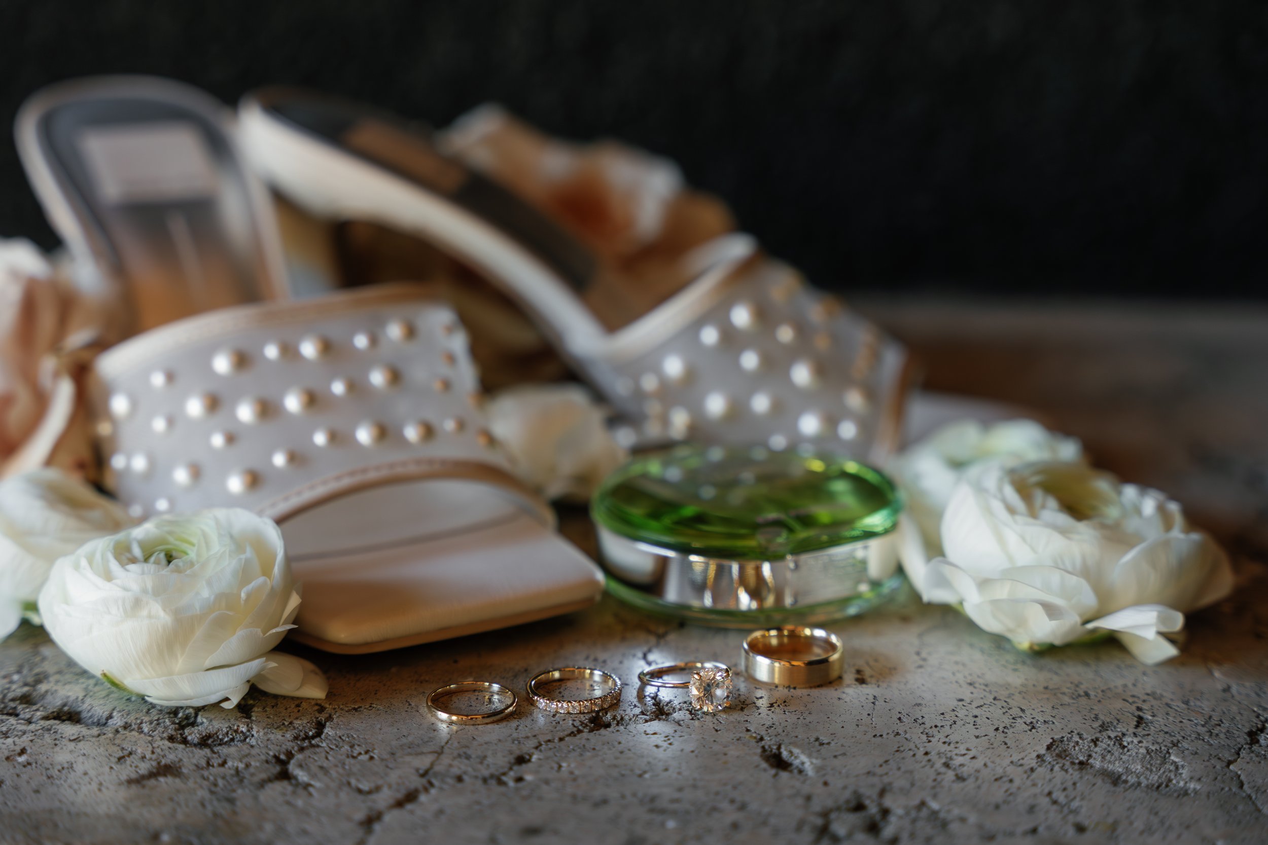 Wedding rings, pearl-adorned shoes, white flowers, and perfume on a textured surface.