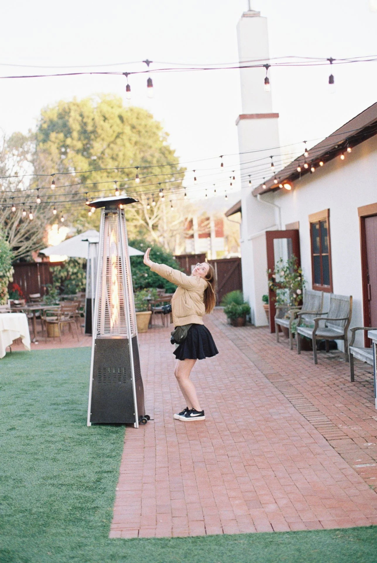 A woman in a beige jacket and black skirt stands next to a patio heater outdoors, reaching out towards the heater with a smile. The setting is a backyard patio with string lights, outdoor furniture, and a brick pathway.