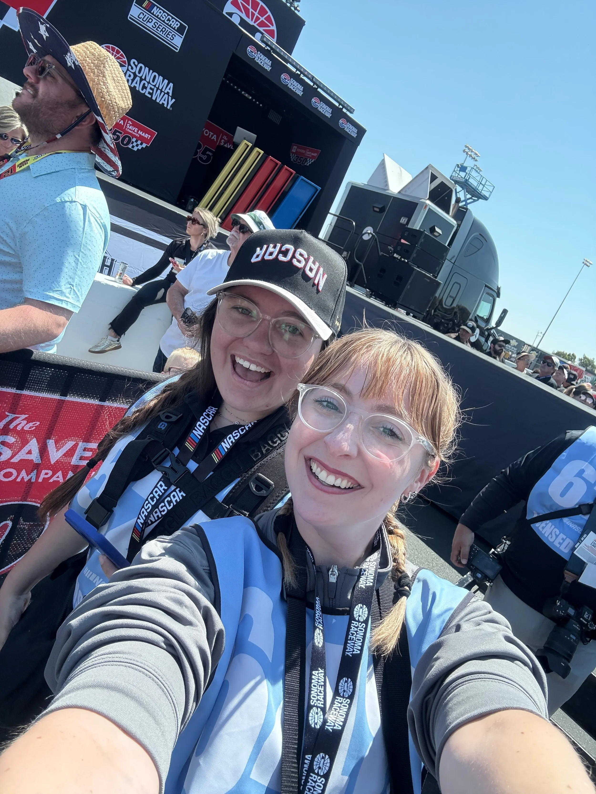 Two women smiling and taking a selfie at Sonoma Raceway during a NASCAR event. They are wearing race officials' vests and lanyards, with race-related bags and caps. In the background, there are spectators, a large race stage with logos, and a black t