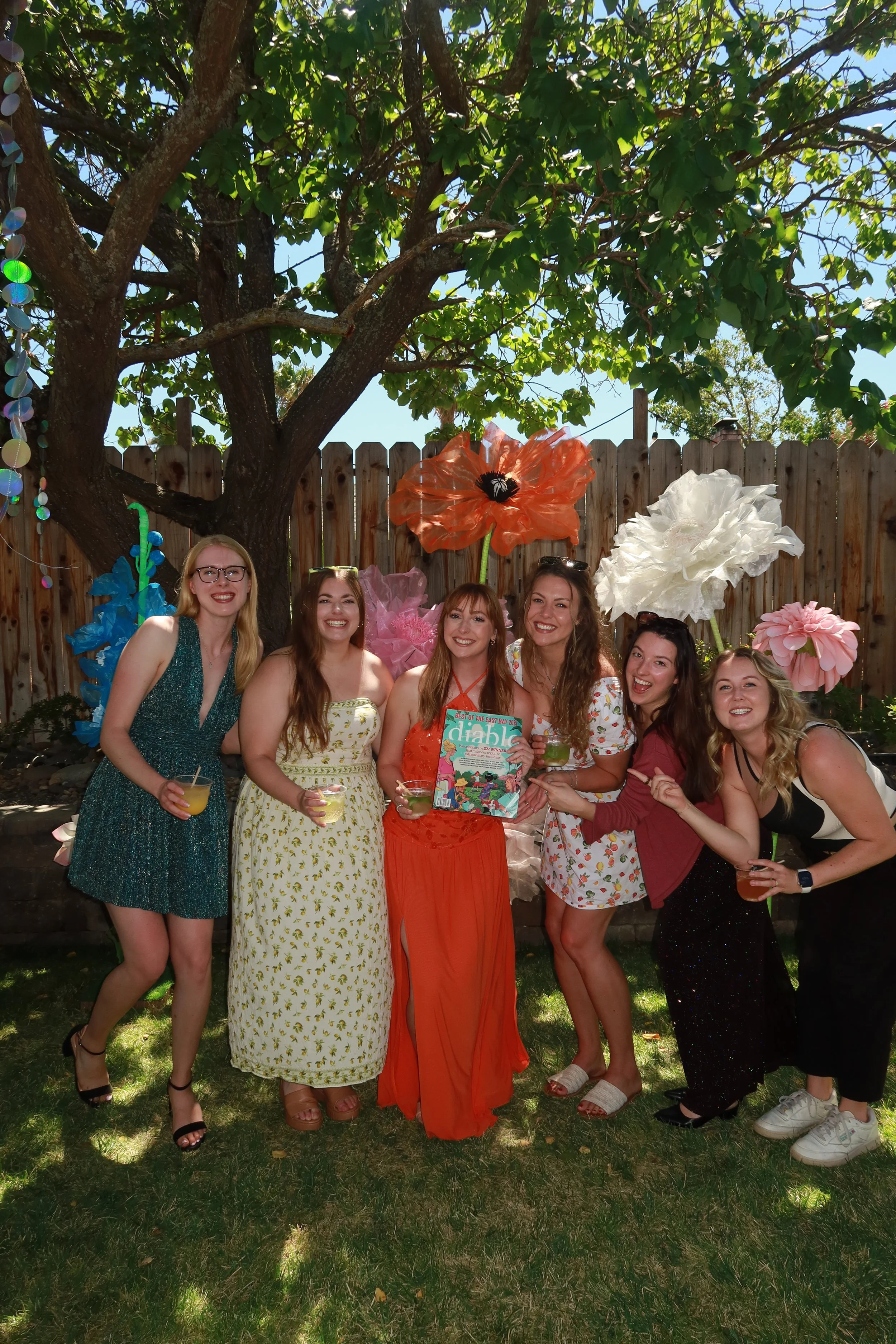 Six women standing together outdoors in front of a wooden fence and decorative paper flowers, smiling and holding drinks, at a garden party or celebration.