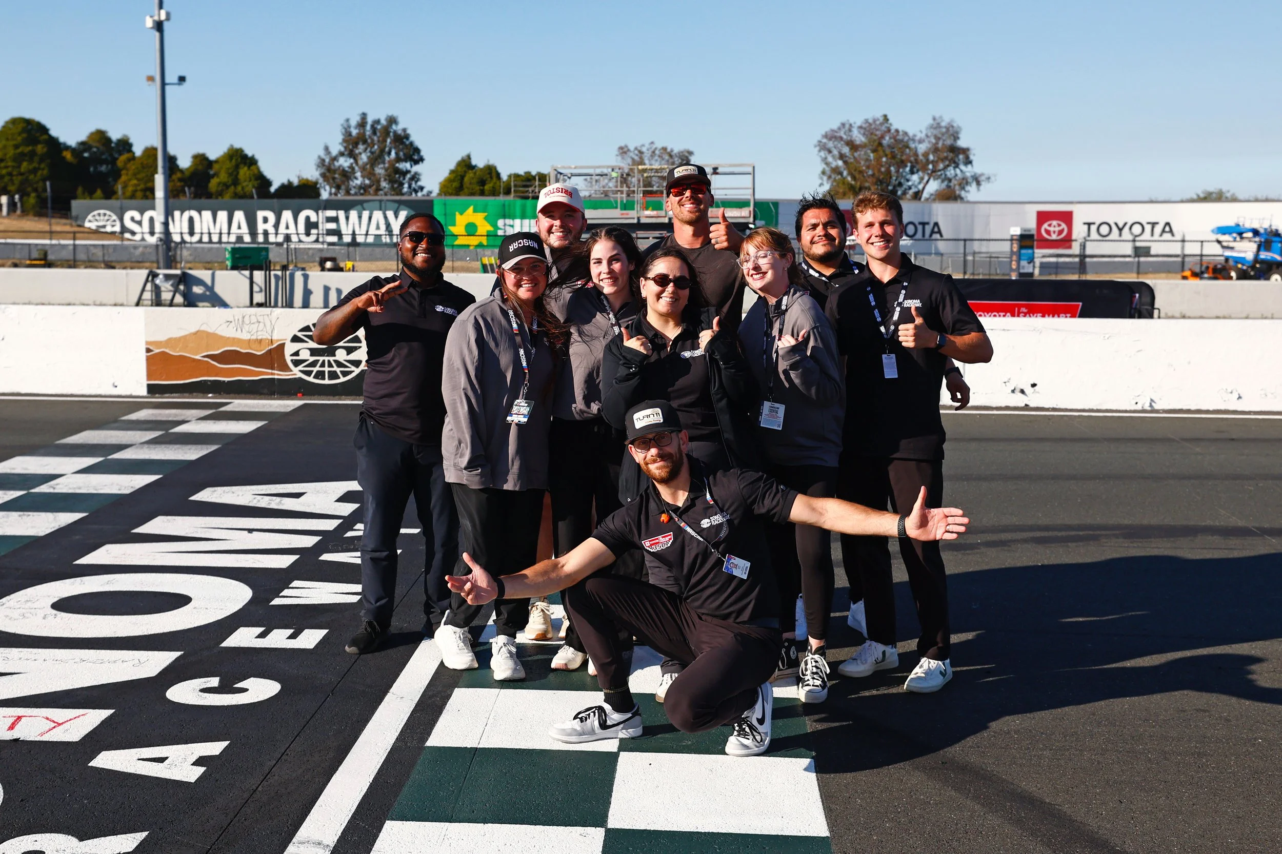 A group of people at a race track posing for a photo, celebrating after a race event at Sonoma Raceway, with some making thumbs-up gestures and smiling.