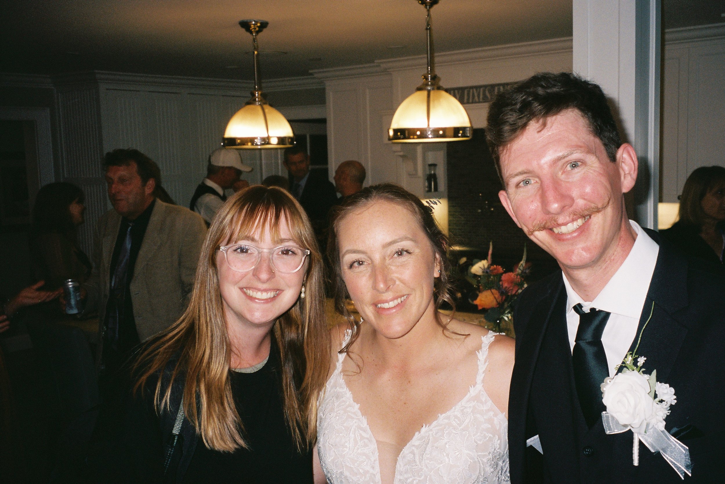 Three people smiling at a social event, with a woman in a white dress in the center and a man in a suit with a white rose boutonniere on the right, and a woman with red hair and glasses on the left, in a warmly lit indoor setting.