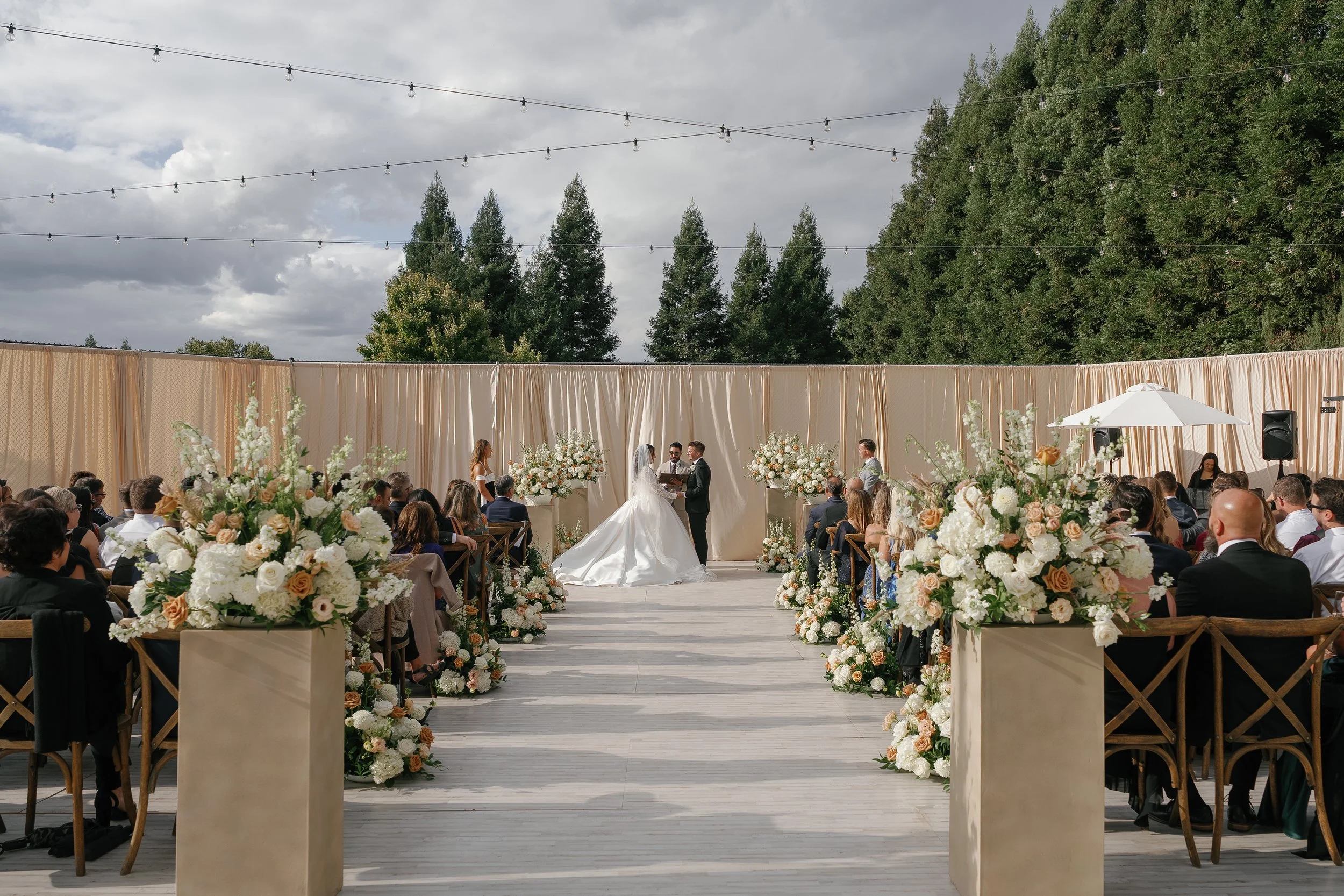 Outdoor wedding ceremony in Chico with a bride and groom exchanging vows, surrounded by floral arrangements and seated guests, under cloudy sky with string lights overhead.