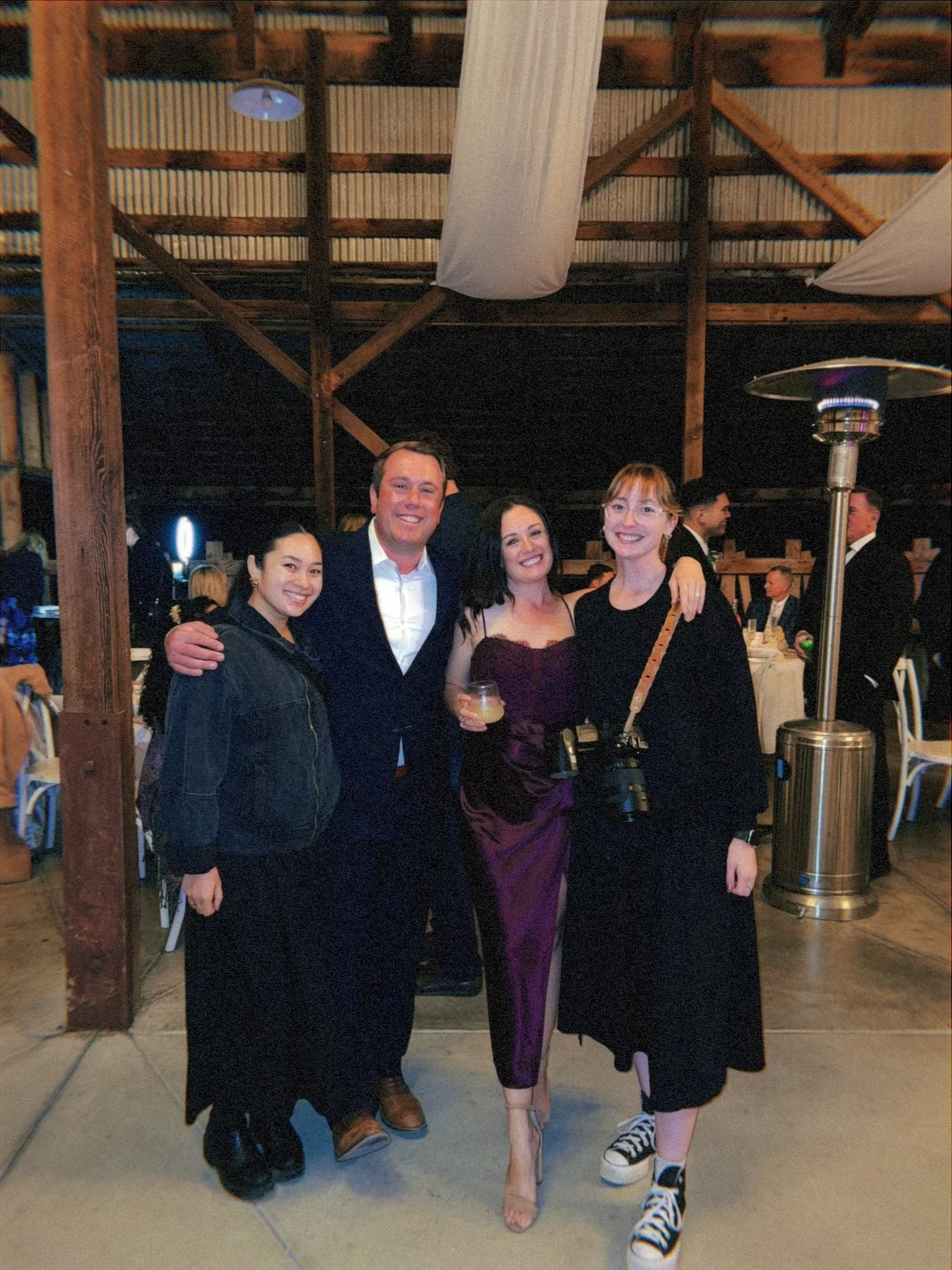 Four people standing together at a social event inside a rustic barn, smiling at the camera. They are dressed semi-formally, with two women in dresses, one in purple and the other in black, and two men in suits. The woman on the right is holding a ca