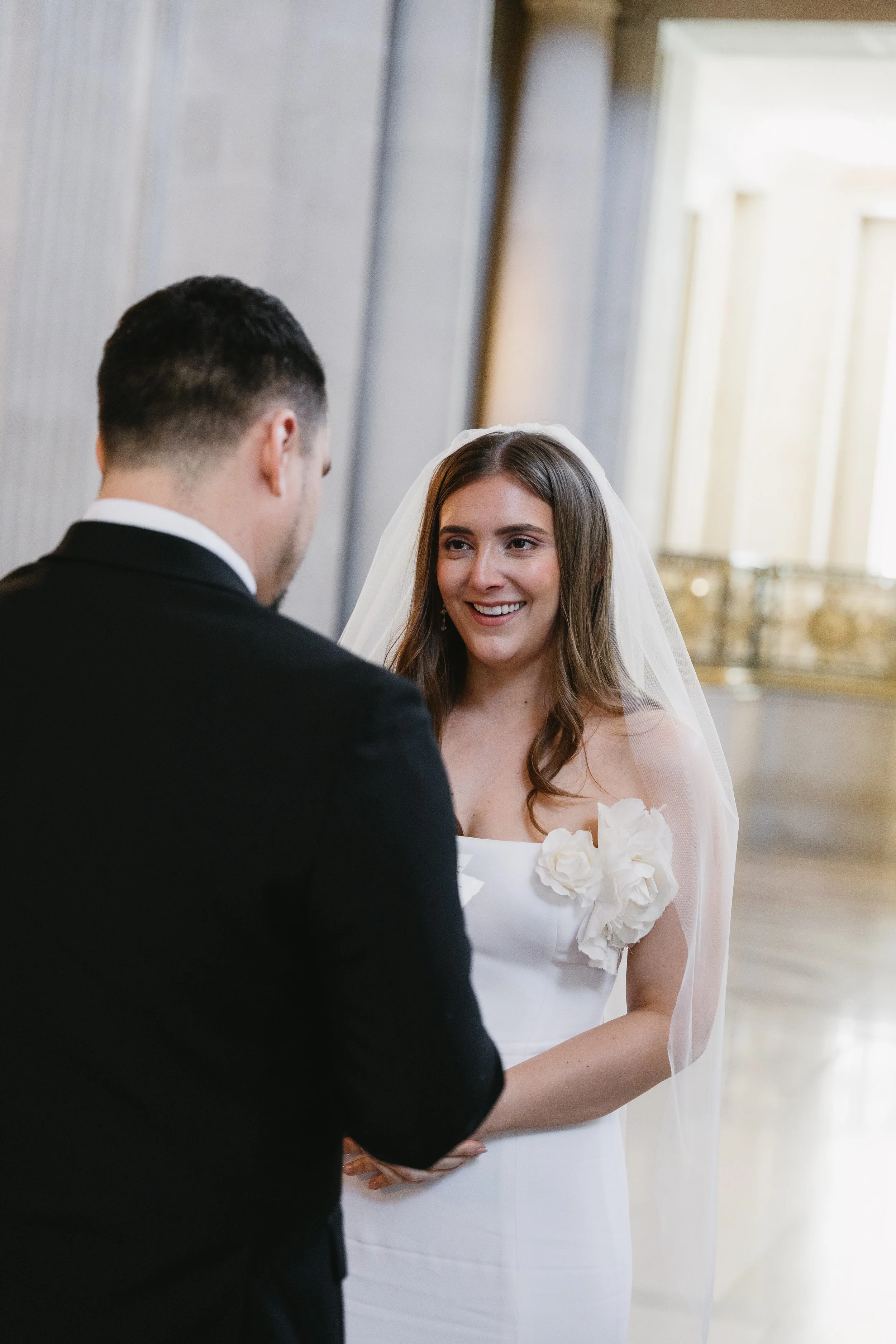 A bride with a veil and white dress smiling at her groom during a wedding ceremony.