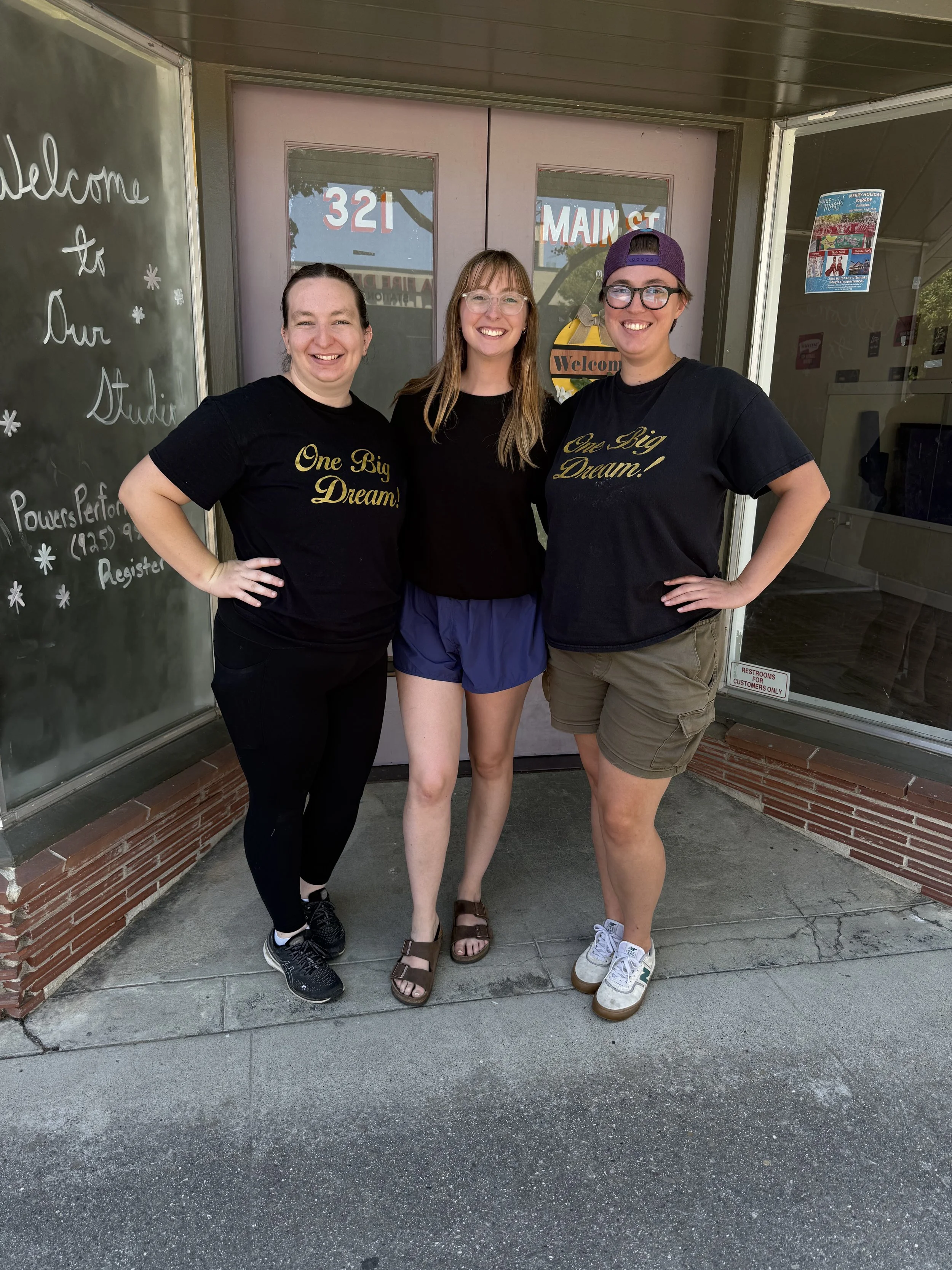 Three women smiling and standing together outside a building with a pink entrance door. Two women are wearing matching black t-shirts that say 'One Big Dream!' and the third woman is in a black shirt with purple shorts. The scene is sunny with reflec