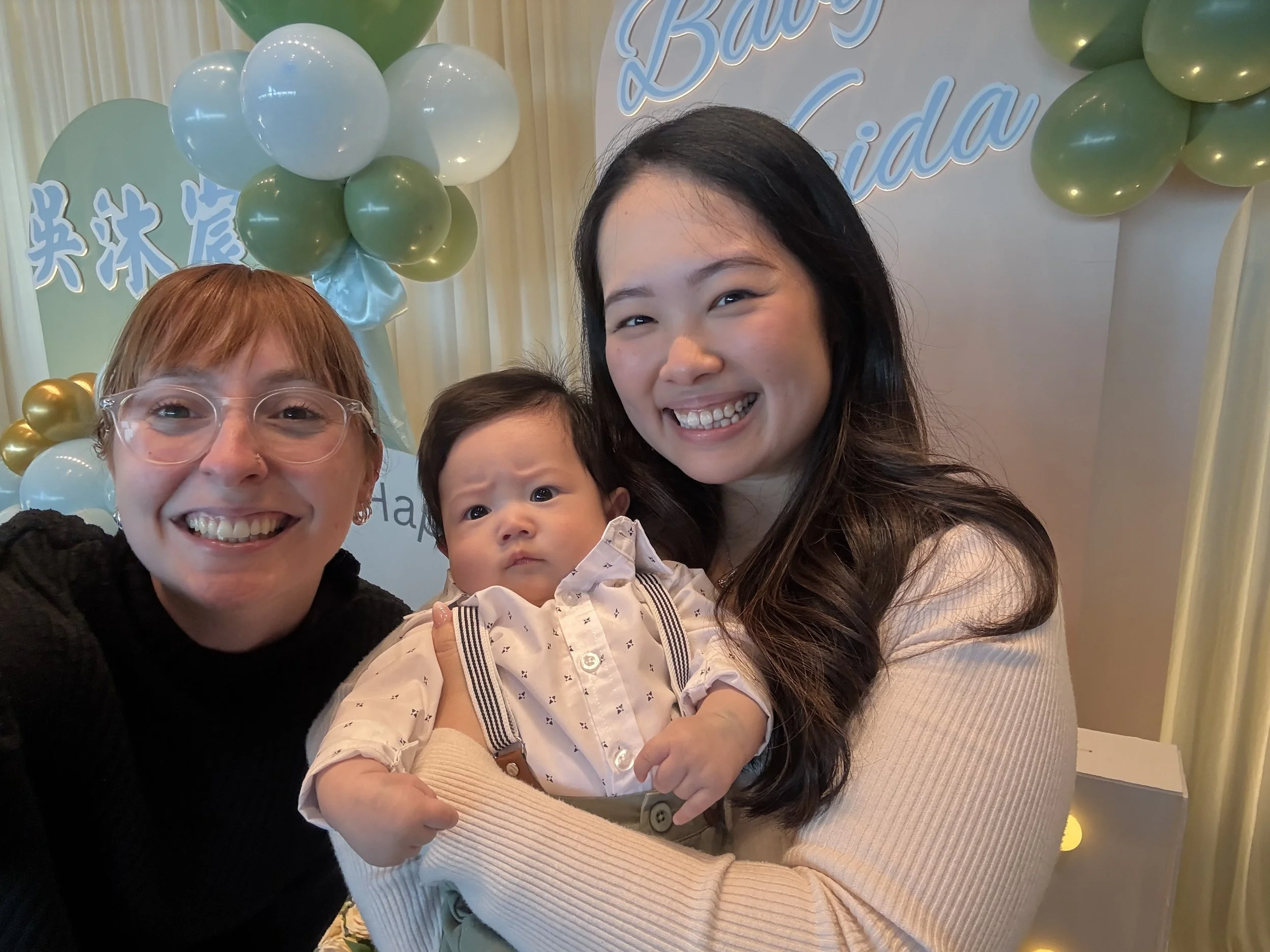 Three smiling women and a baby pose for a photo at a celebration. In the background, balloons and a sign with the words 'Baby' and 'Laida' are visible.