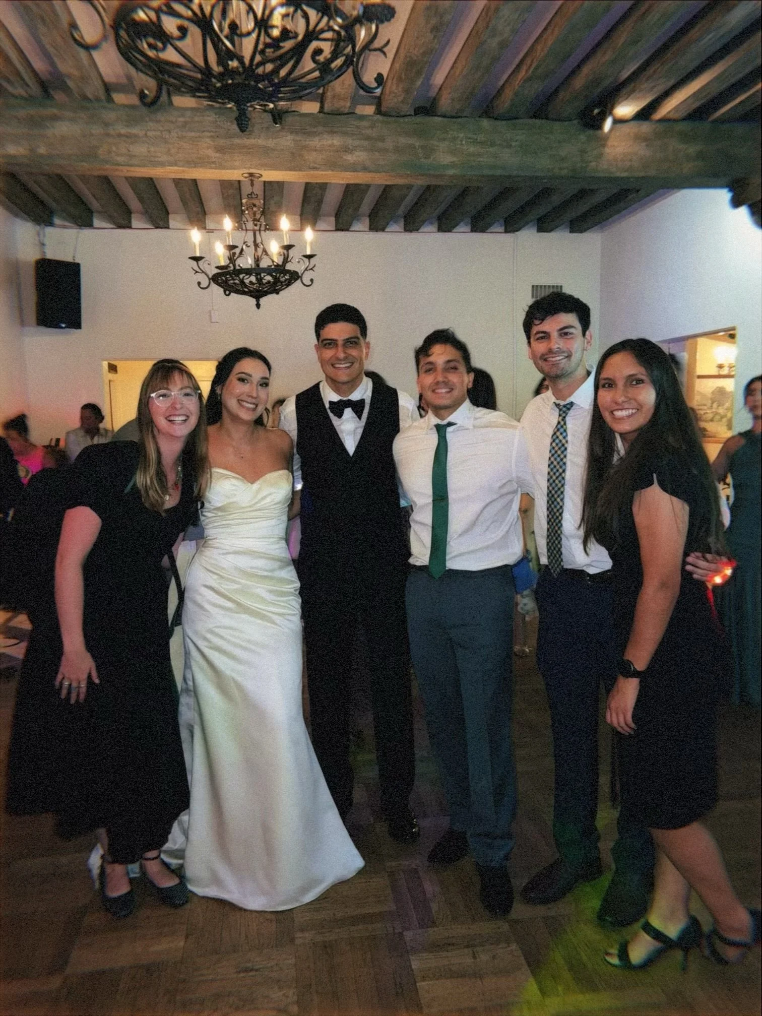 A group of six people, dressed in formal attire, standing together indoors with a chandelier hanging from a rustic wooden ceiling. They are smiling and appear to be celebrating a special occasion, possibly a wedding.