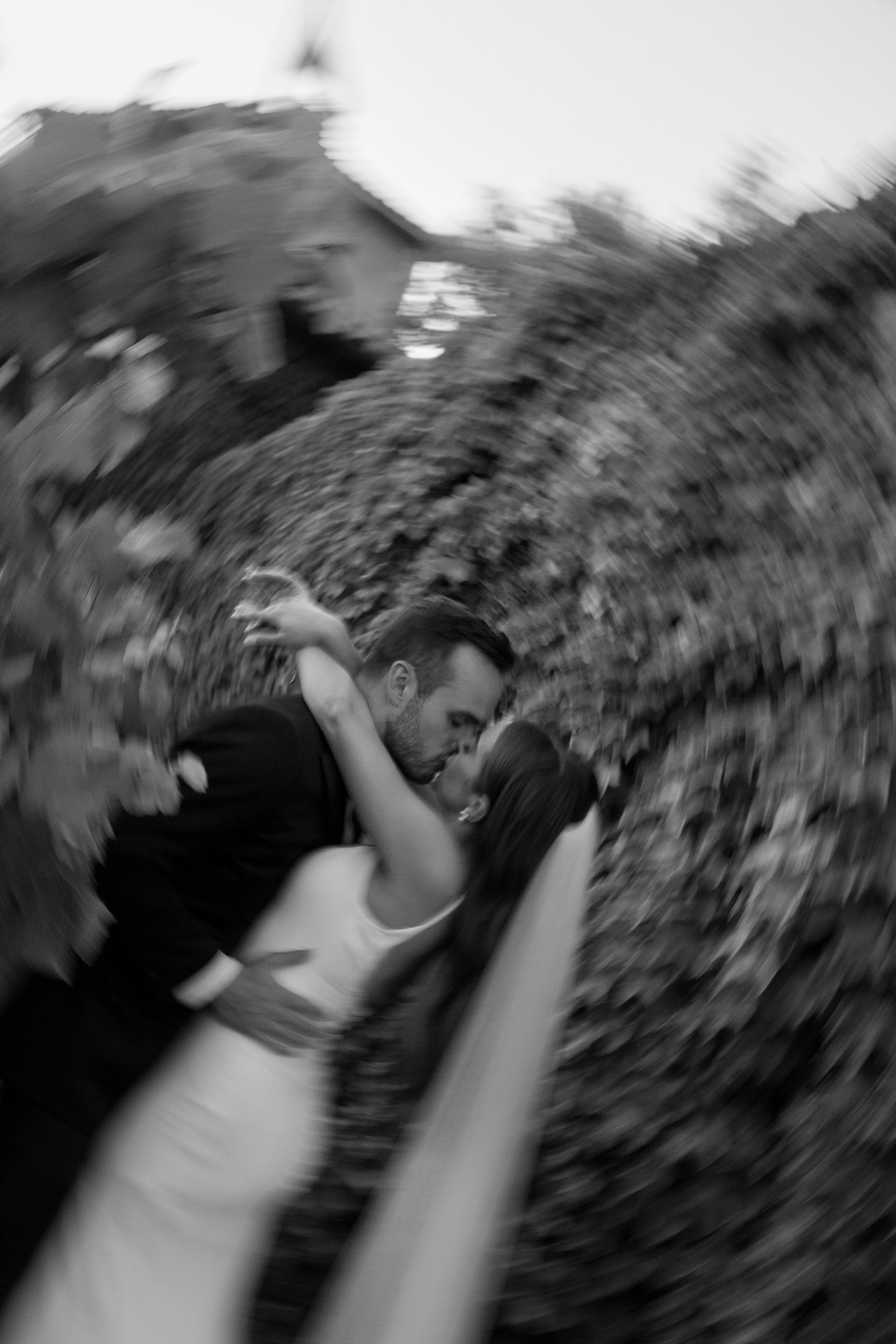 A black and white photo of a couple kissing outdoors, with motion blur creating a swirling effect around them.