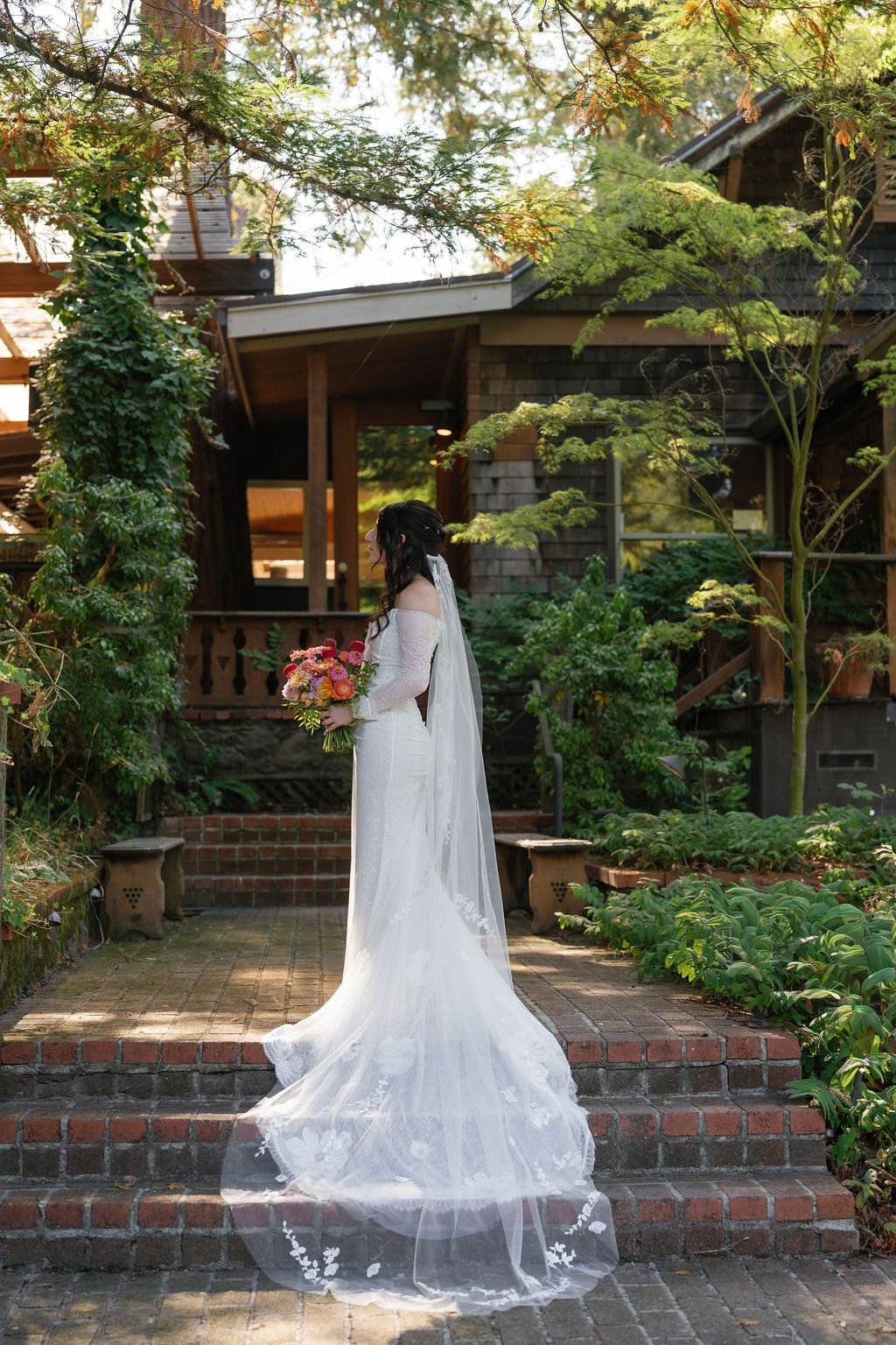 A bride in a white wedding dress holding a bouquet of colorful flowers stands on brick steps outdoors, surrounded by lush greenery and trees, in front of a rustic wooden house.