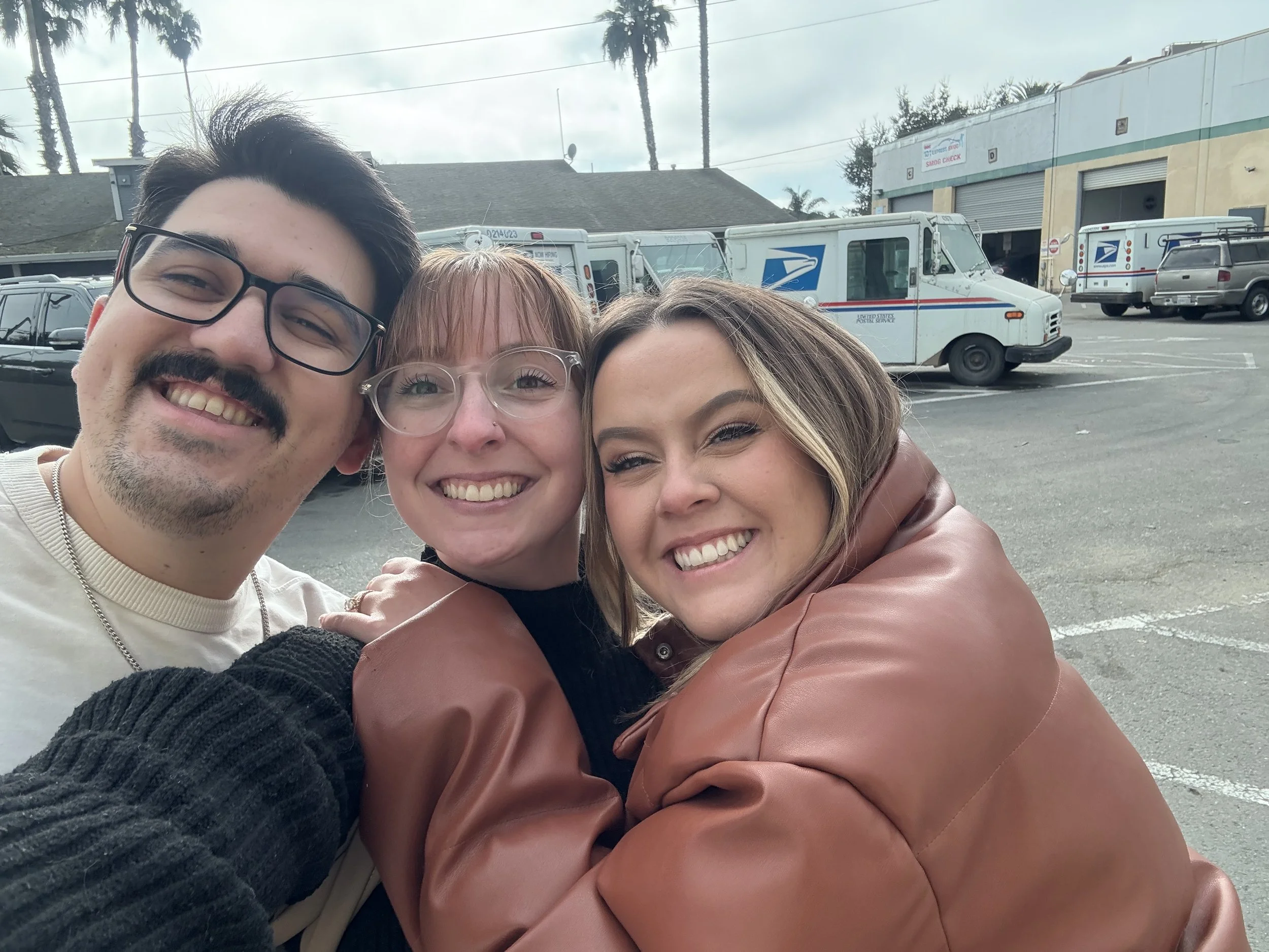 Three people smiling and taking a selfie outdoors with postal service trucks and cars in the background.