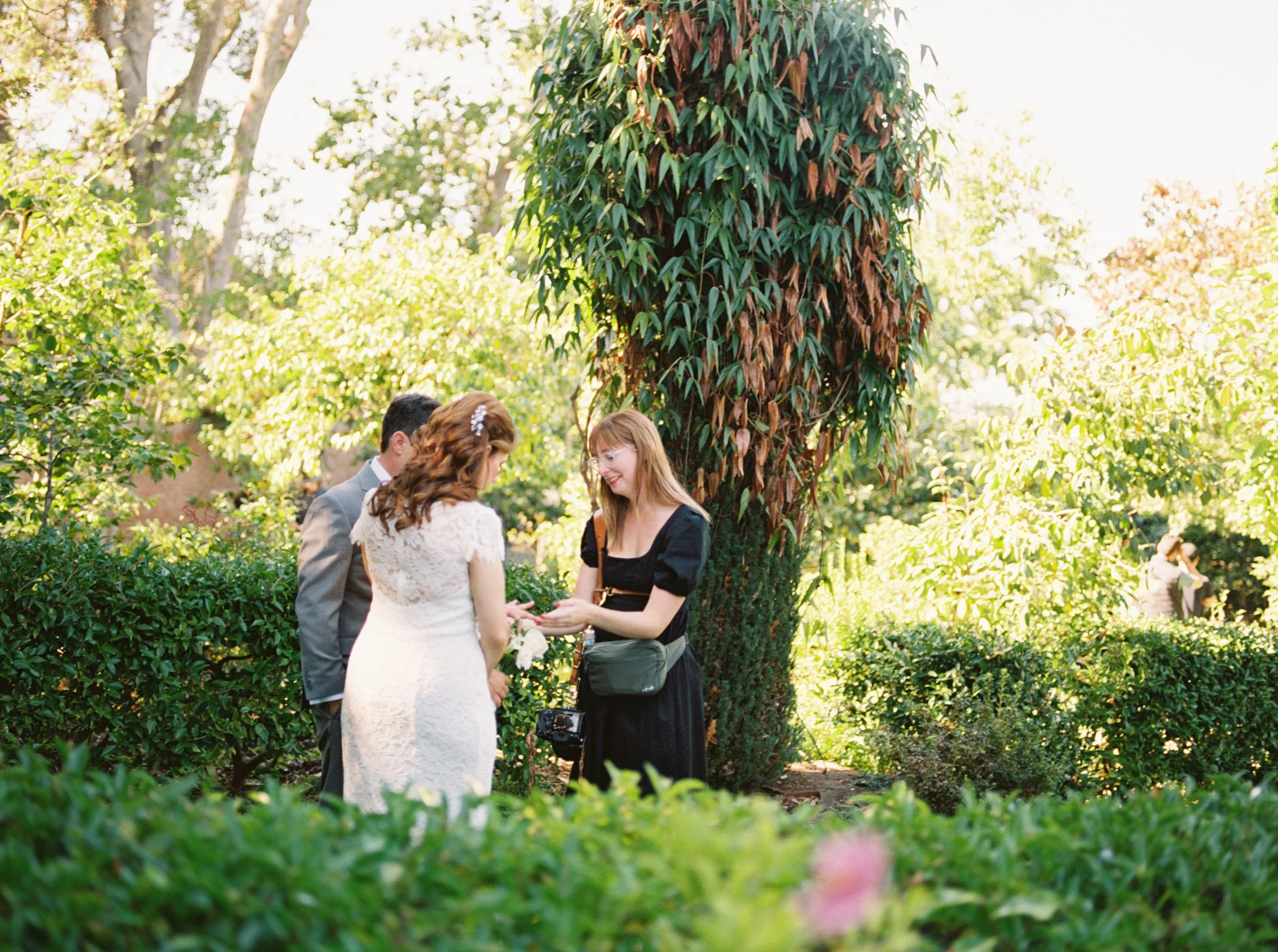 A bride and groom exchanging vows outdoors with a woman officiant in a garden setting.