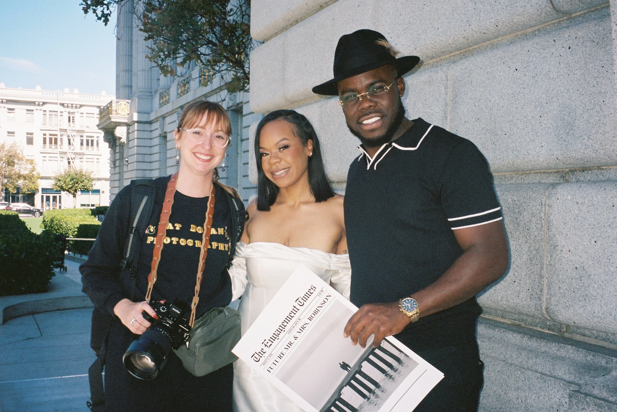 Three people posing outdoors, one holding a camera, and a newspaper at san Francisco city hall taking engagement photos