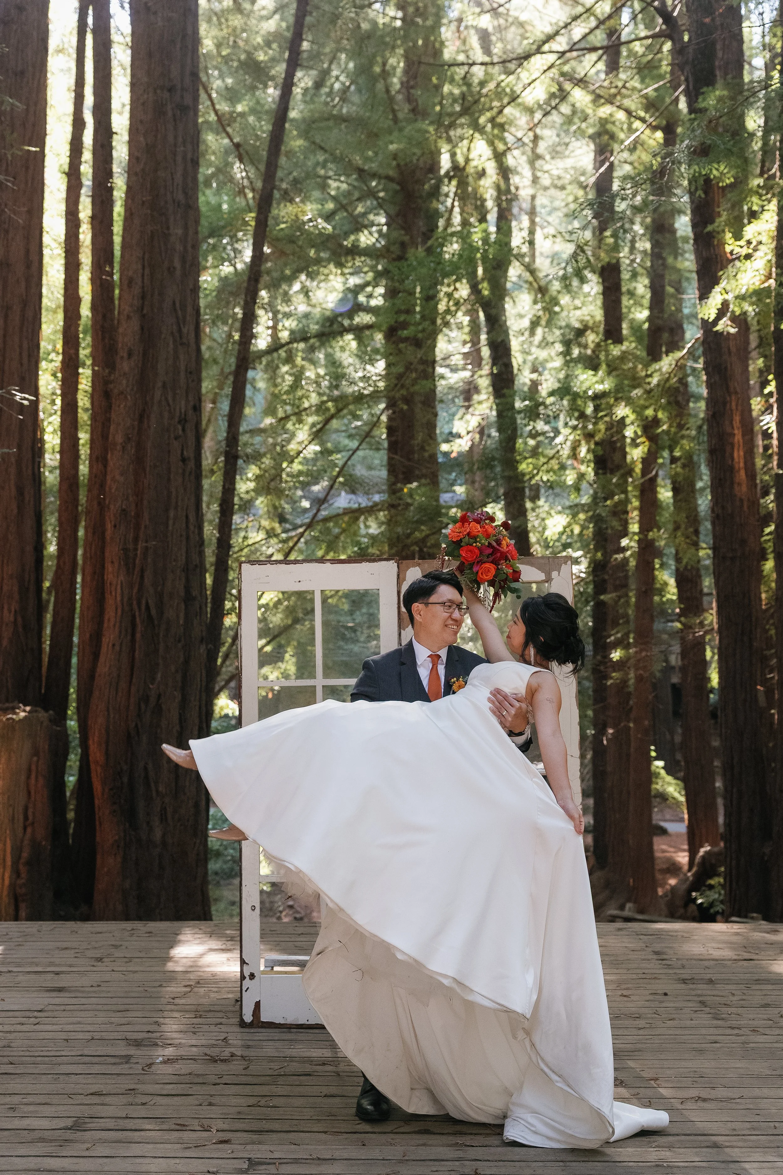 A wedding couple is posing outdoors in a forest. The bride, in a white wedding gown, is being carried by the groom, who is dressed in a suit. She is holding a bouquet of red and orange flowers, and they are smiling at each other. There is a window frame behind them, and the scene is illuminated by natural sunlight filtering through the tall trees.