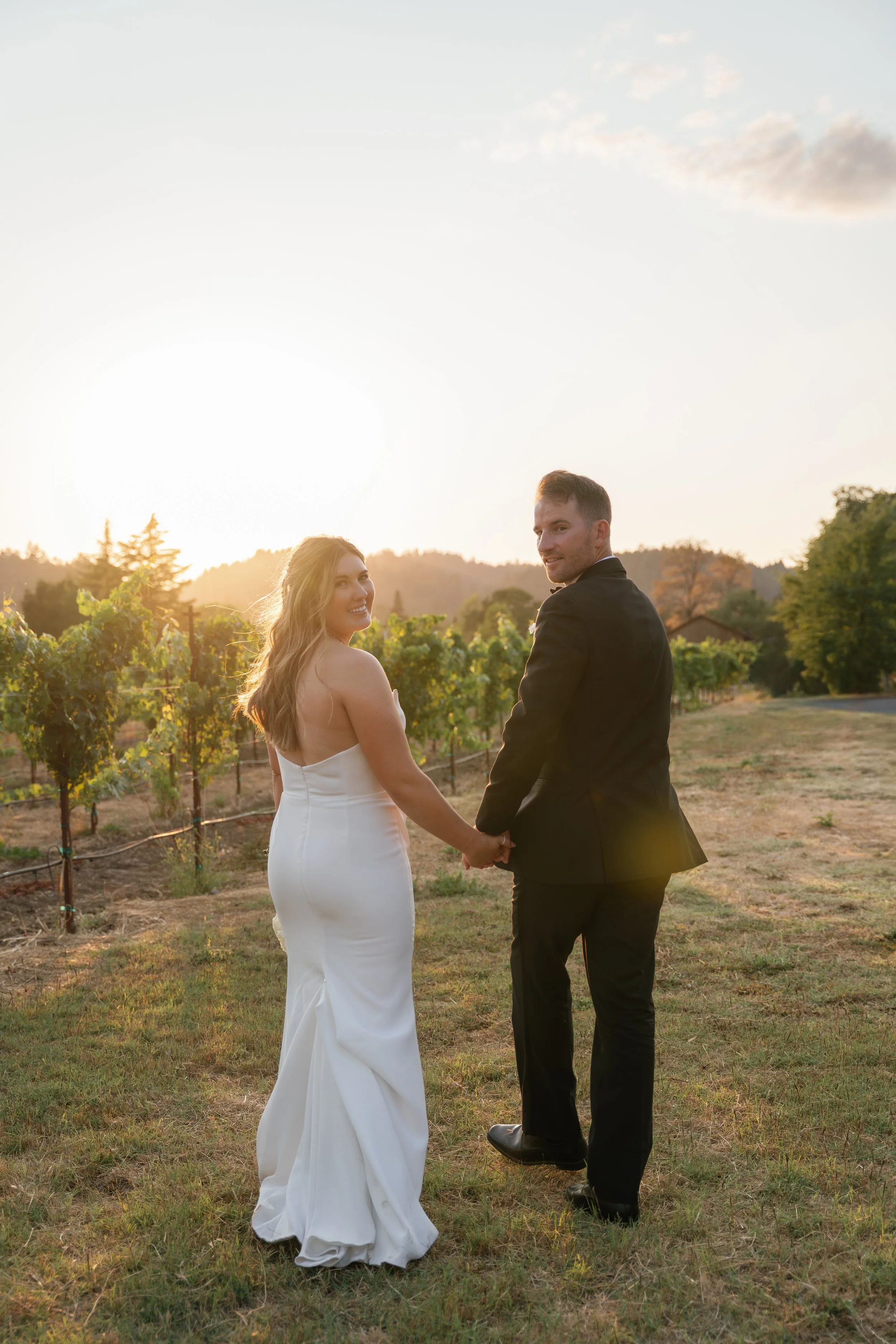 A newlywed couple holding hands and looking back at the camera in a vineyard during sunset.