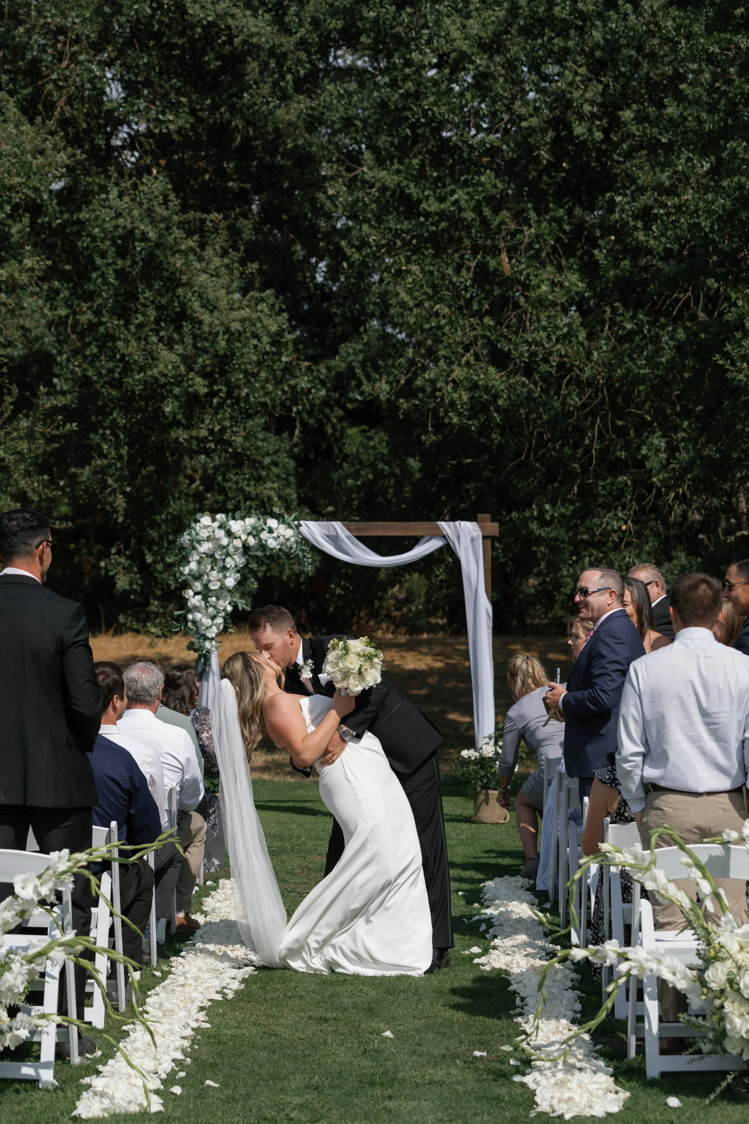 A newlywed couple shares a kiss at an outdoor wedding ceremony, with guests seated on either side of a petal-lined aisle. The bride holds a bouquet of white flowers and wears a white gown with a veil, while the groom is dressed in a black tuxedo. The