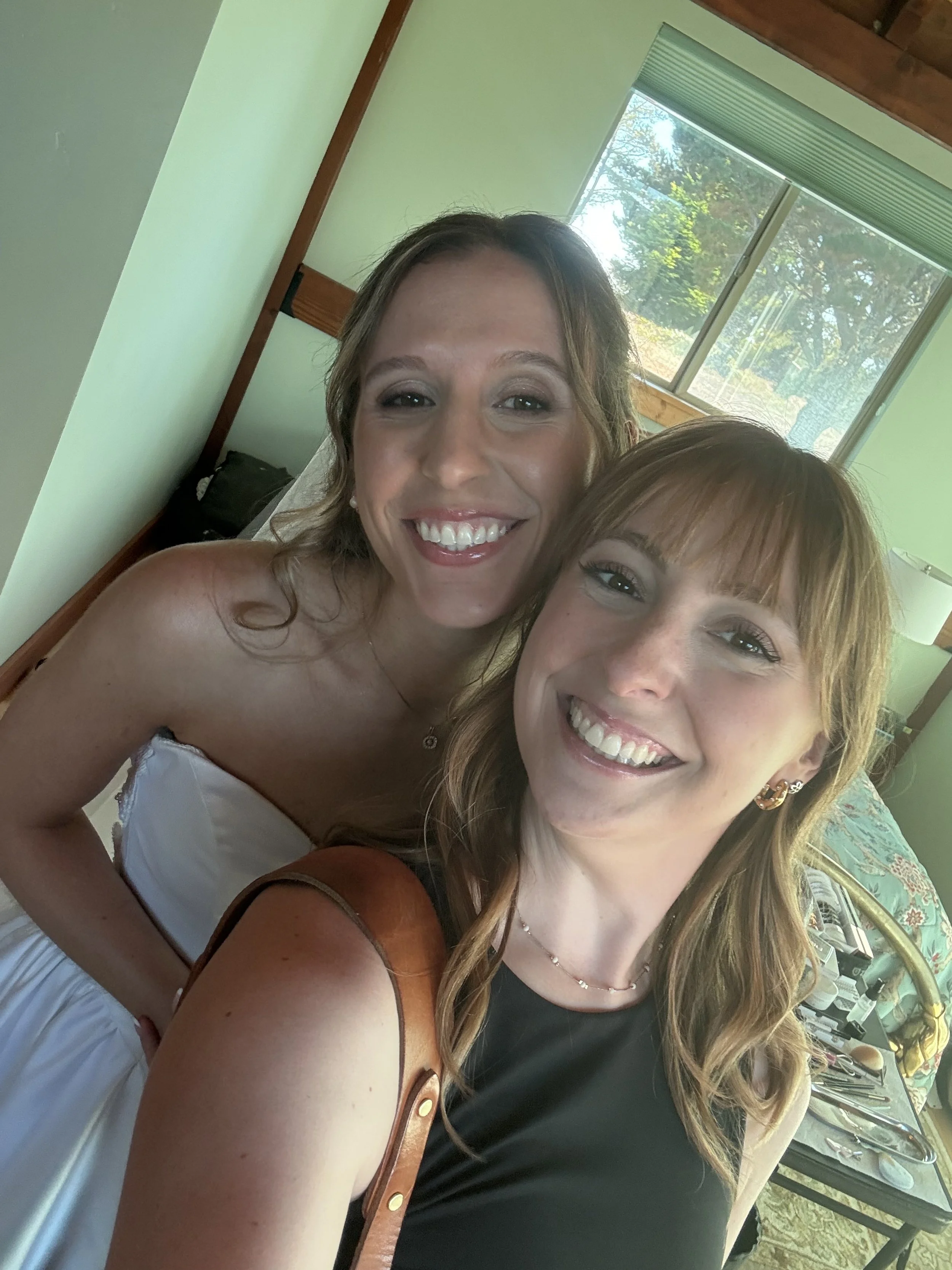 Two women smiling and taking a selfie indoors near a window.