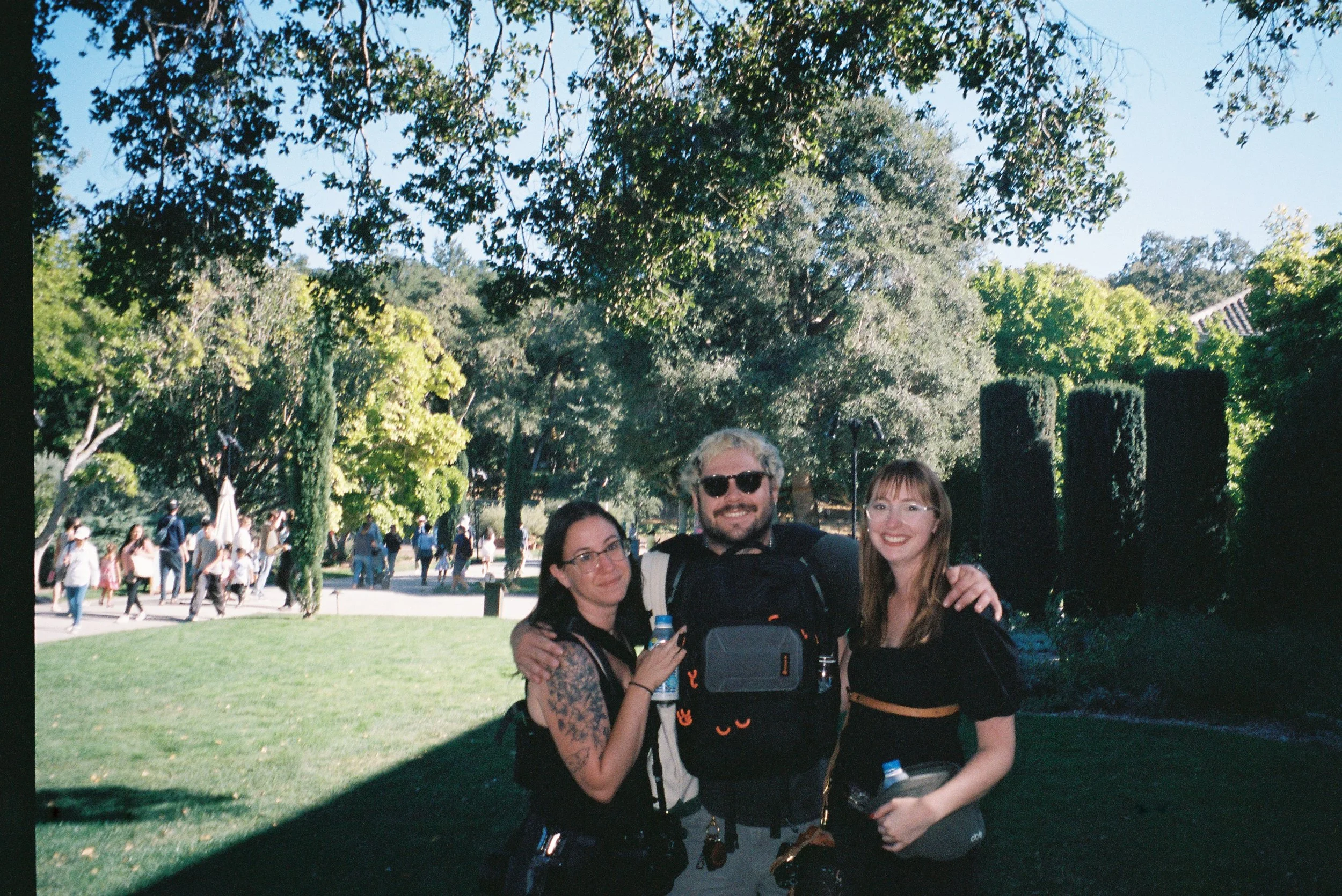 Three friends smiling and hugging outdoors in a park with green trees and people walking behind them.