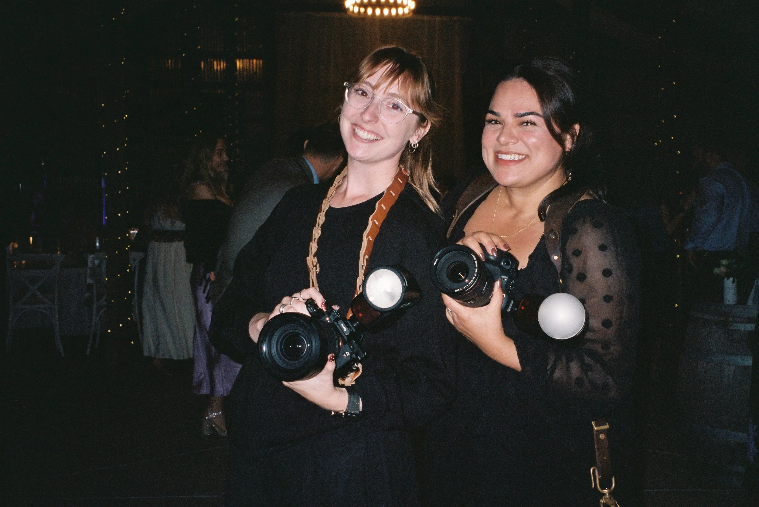 Two women smiling and holding cameras at a social event indoors, with people and dim lighting in the background.