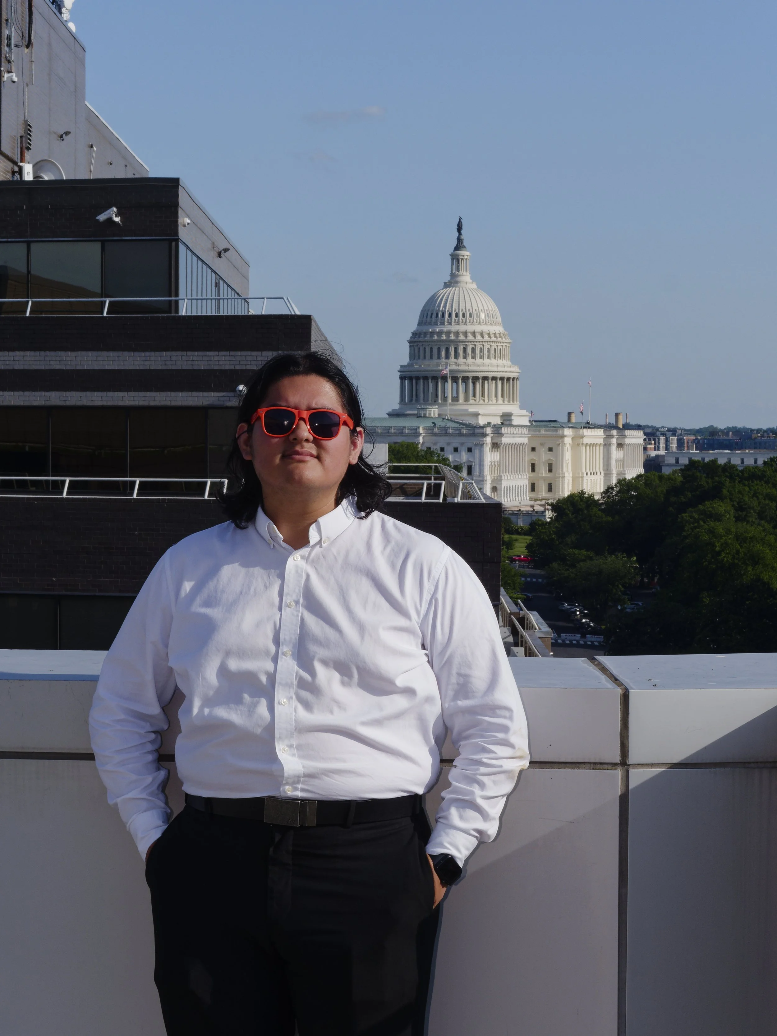 A person wearing a white shirt and red sunglasses stands on a rooftop with the U.S. Capitol building in the background.