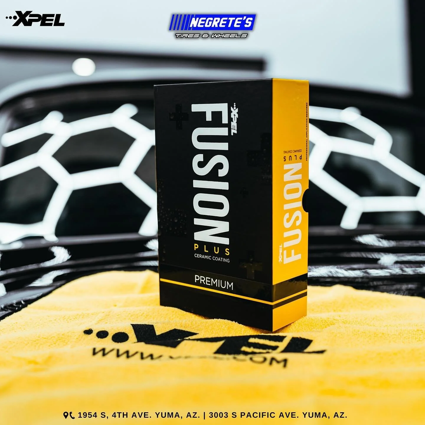 Box of Fuse Plus ceramic coating product placed on a yellow cloth with black text, in front of a car grille with a honeycomb pattern, with promotional stickers in the background.