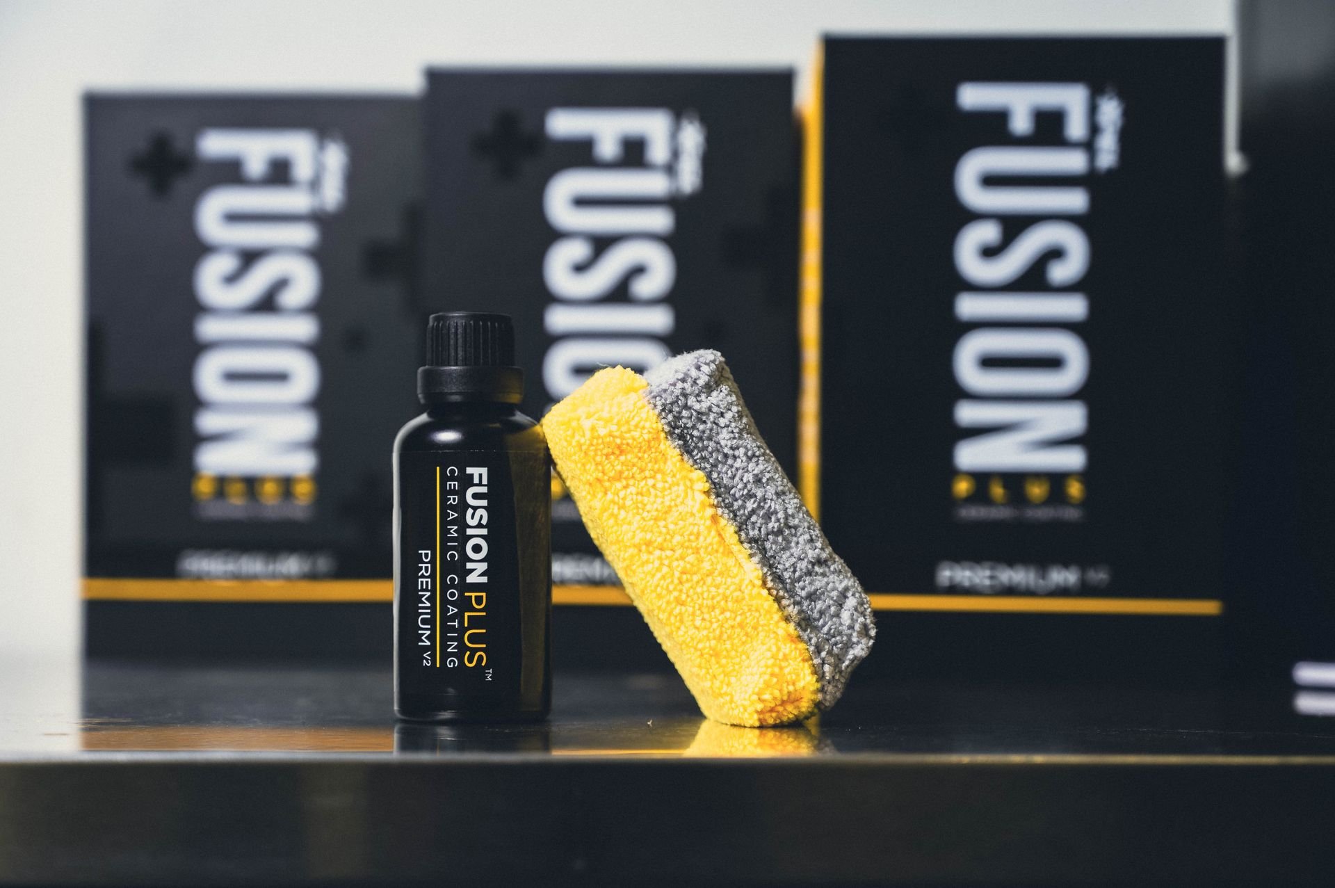 A black bottle labeled Xpel Fusion Plus Ceramic Coating Premium V2 with a yellow and gray microfiber applicator pad leaning against it, set against Fusion Plus product boxes in the background.