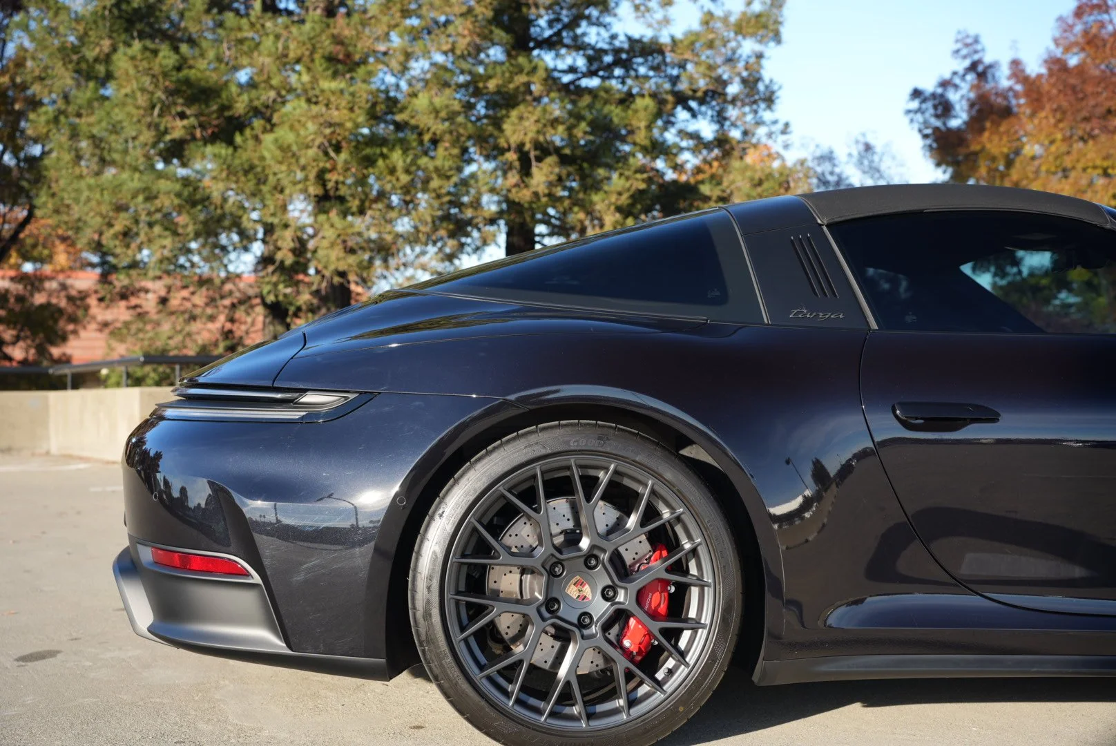 Front side of a black Porsche Targa sports car parked outdoors, with trees and a building in the background, showing sleek curves and alloy wheels with red brake calipers.