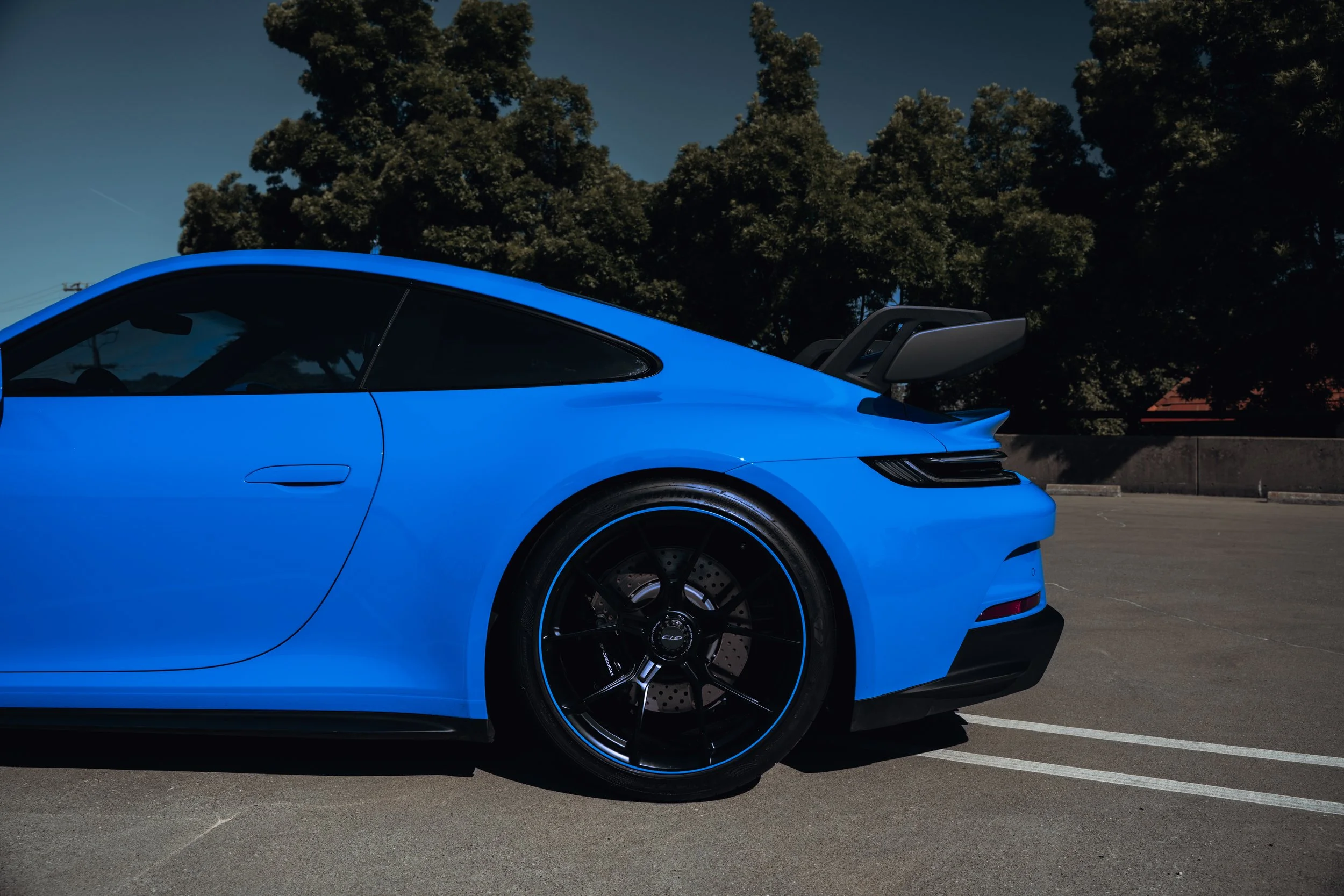 Side view of a bright blue sports car with black wheels and a rear spoiler, parked in an empty parking lot with trees in the background.