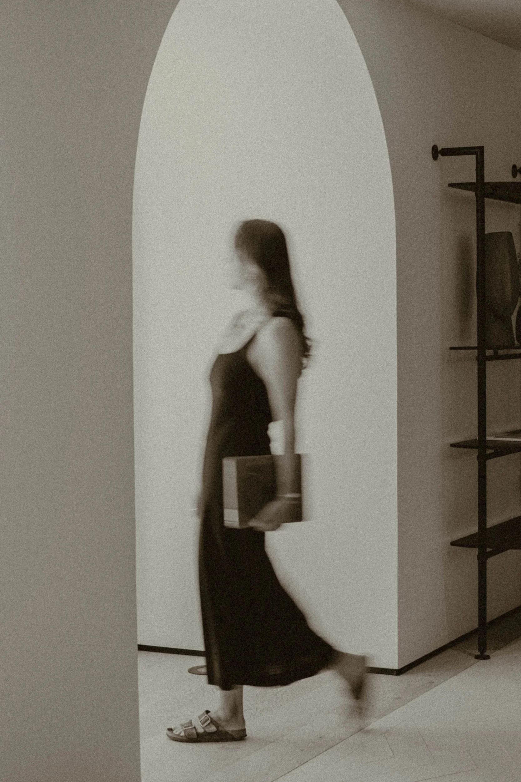 A woman in a black dress and sandals walking with a bag in her hand, captured in motion, inside a building with a curved archway and shelving on the wall.