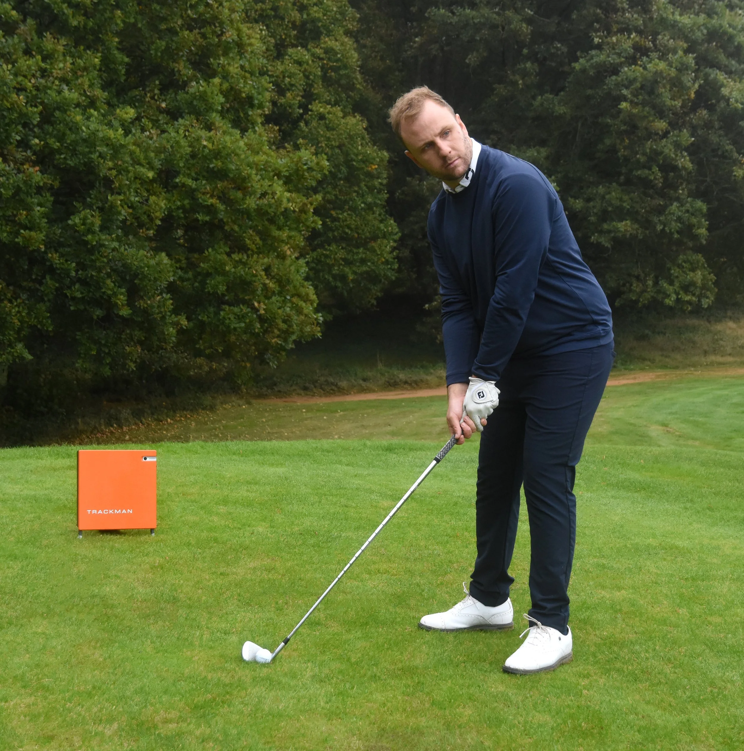 Golfer preparing to swing on golf course with Trackman device nearby.