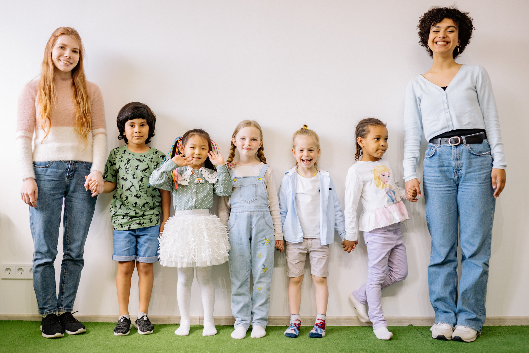 A group of eight diverse kids and two women standing in a row against a white wall, holding hands, smiling, and looking at the camera.