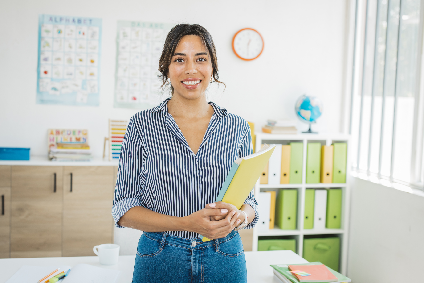 A woman smiling and holding a yellow book in a classroom or office with colorful storage bins, posters, and a globe in the background.