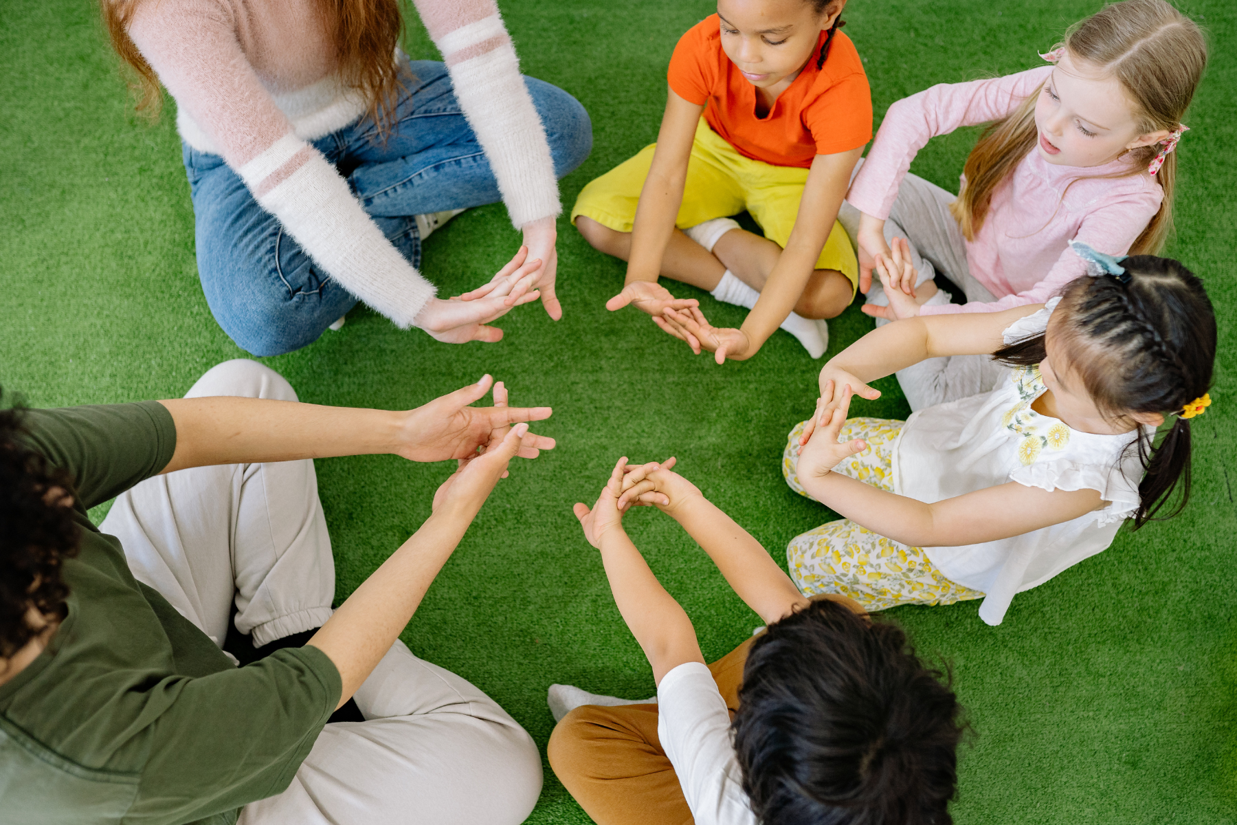 Children and adults sitting on green carpet, engaging in a group activity or game, touching hands in the center.