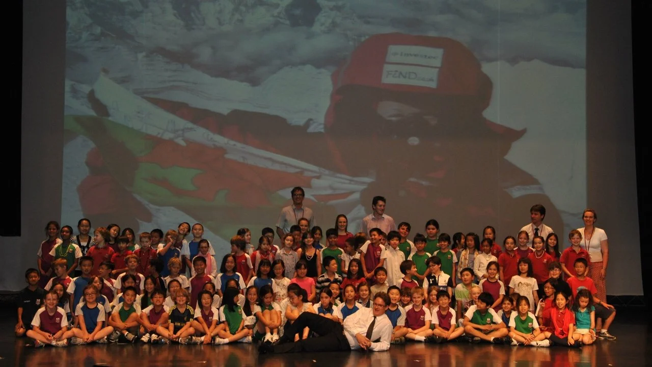 Tori James stands on stage with students at Dulwich College Shanghai infront of a presentation showing an image of Tori James holding a welsh flag standing on Mount Everest