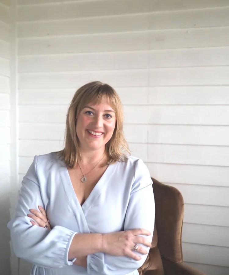 Woman with blonde hair smiling and crossing arms in front of a white shiplap wall, wearing a light blue blouse and a necklace.
