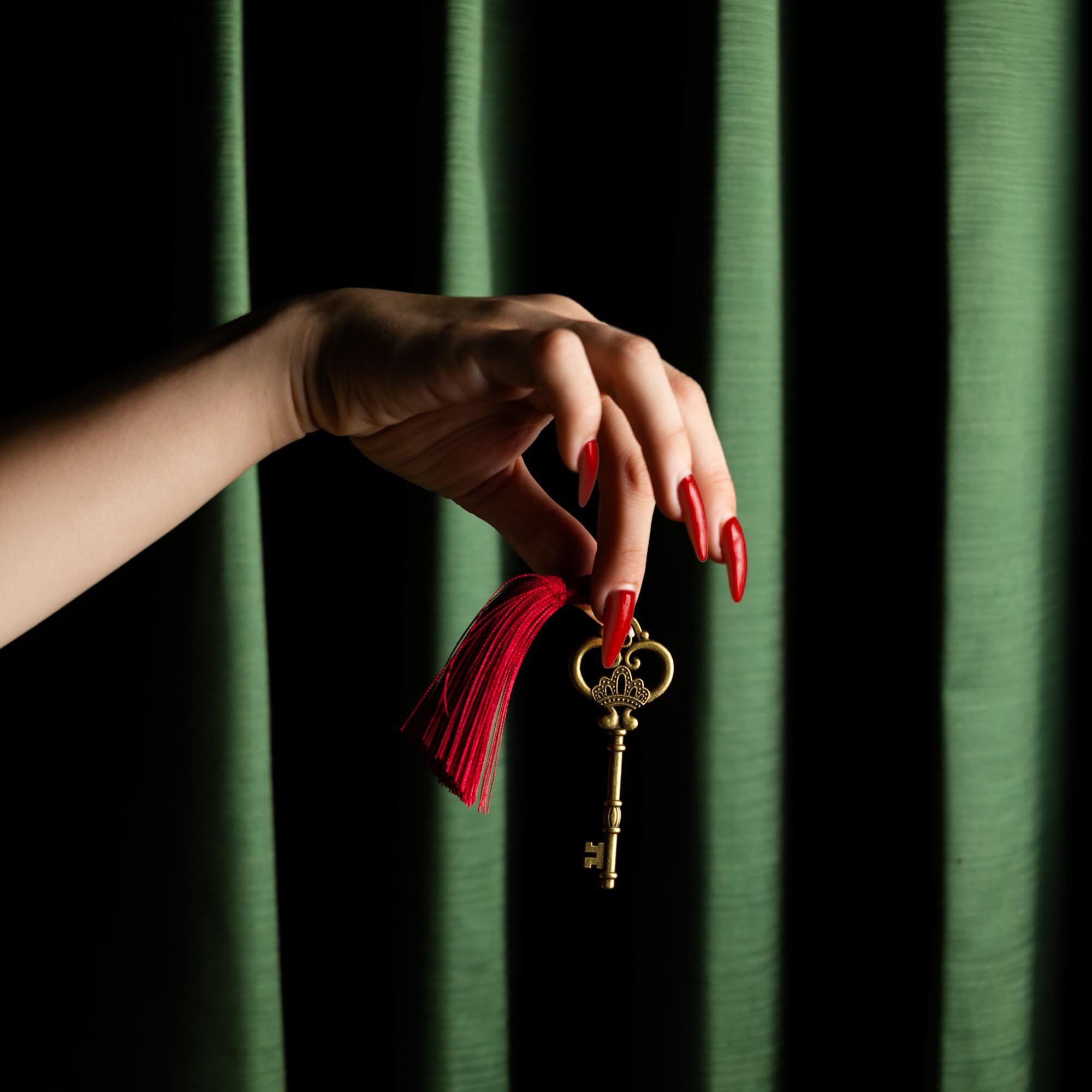 Hand with red nails holding a key with a decorative crown-shaped pendant against a dark background with green-striped curtains.