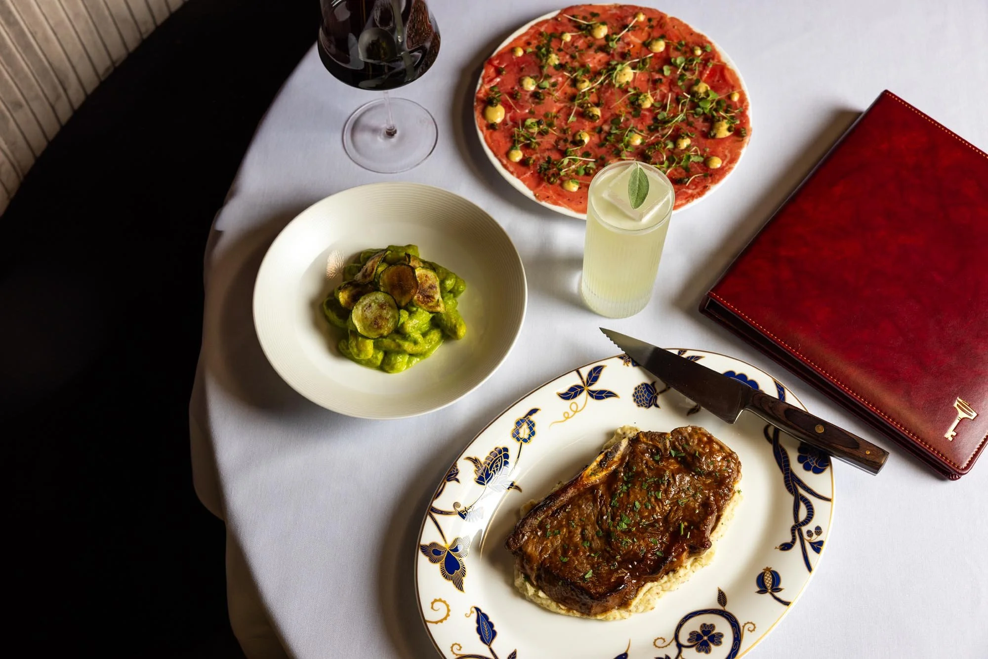 A table set with Il Segreto's signature offerings,  a glass of red wine, a glass of pale yellow beverage with a leaf garnish, and a wine book, all on a white tablecloth.