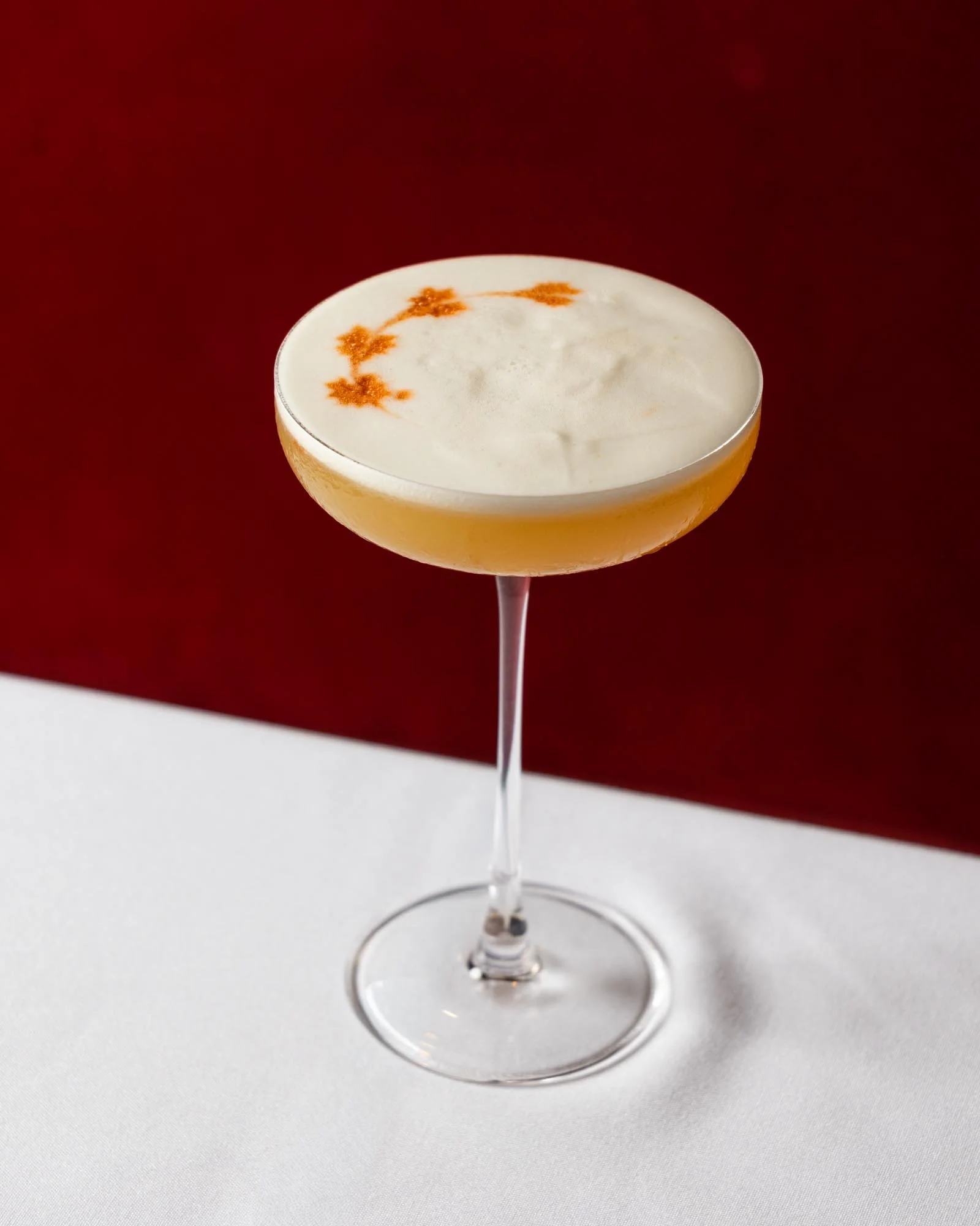 A cocktail glass with a frothy, light-colored beverage topped with orange spice and cinnamon, set against a red and white background.
