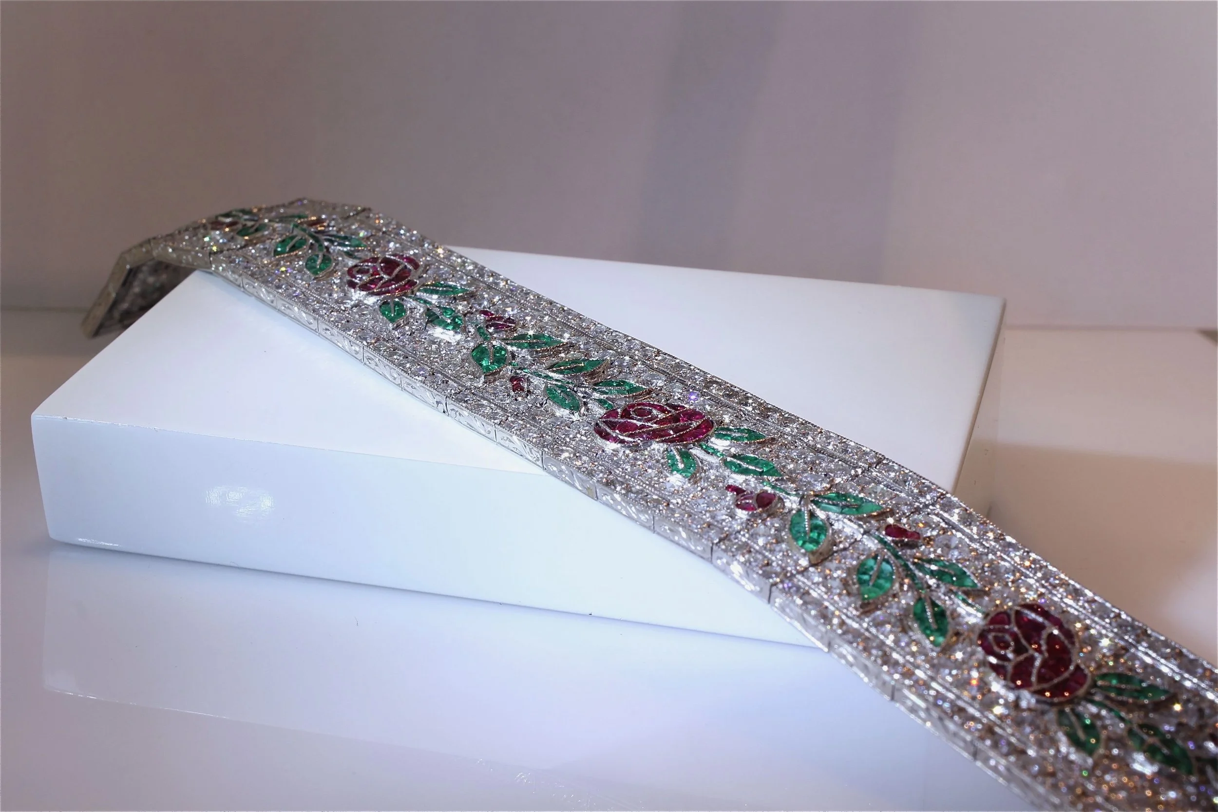 A decorative silver bracelet with engraved floral patterns, embedded with red and green stones, displayed on a white surface.