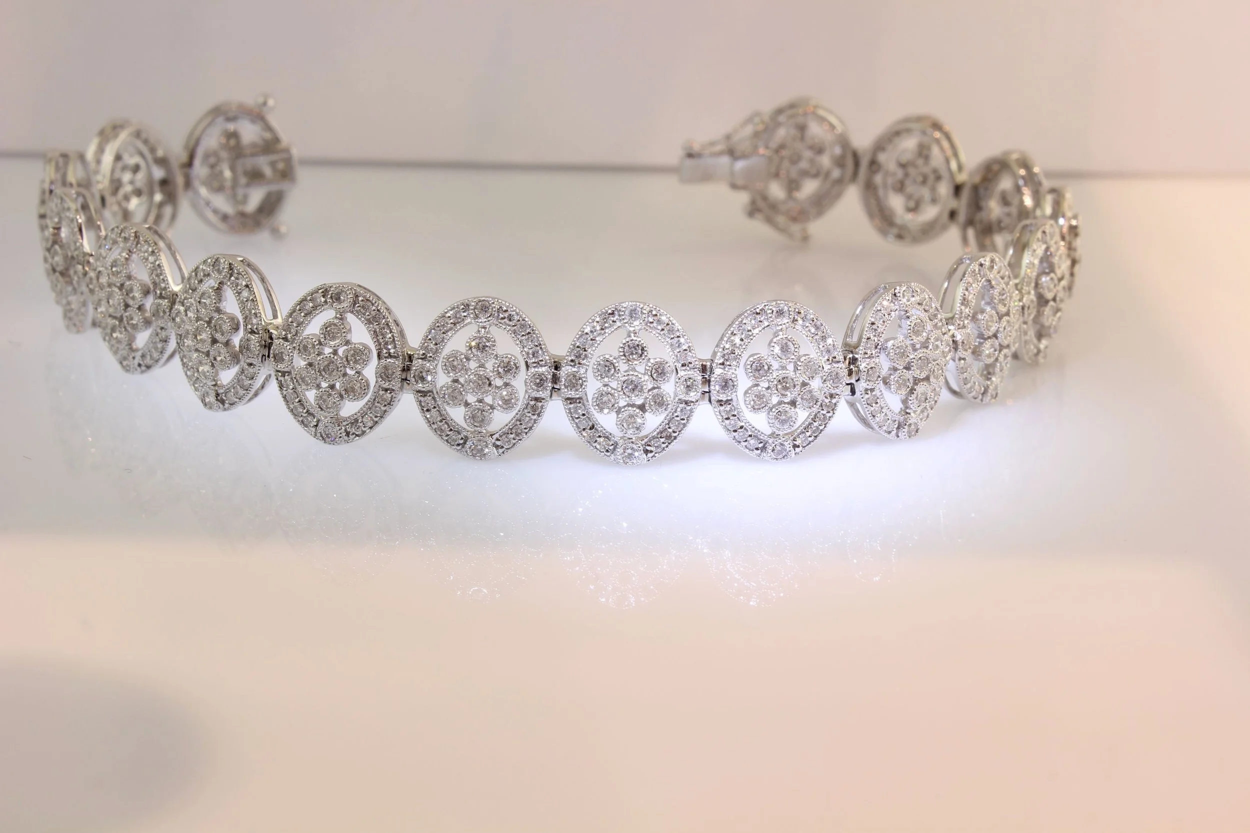 A silver bracelet with intricate oval-shaped designs encrusted with small diamonds, placed on a reflective surface.
